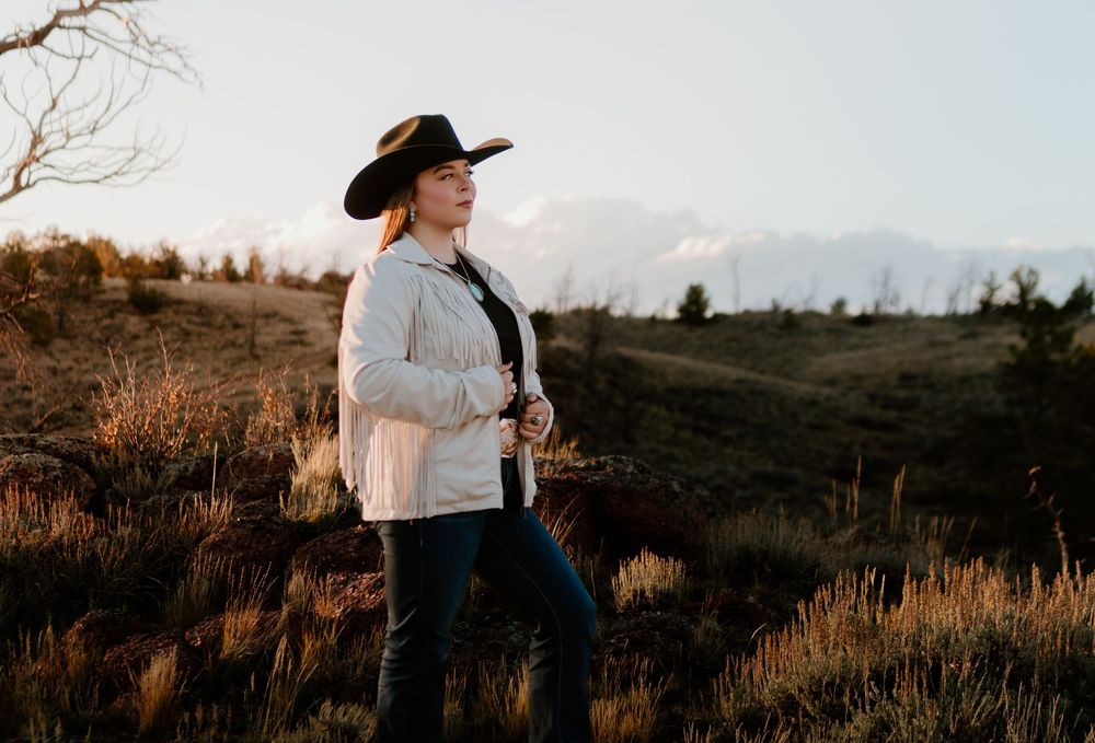 Woman in cowboy hat and white jacket stands in golden field at sunset.