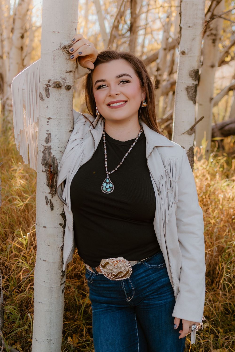 Young woman in white fringe jacket, turquoise necklace, and western belt buckle posing among aspen trees outdoors.