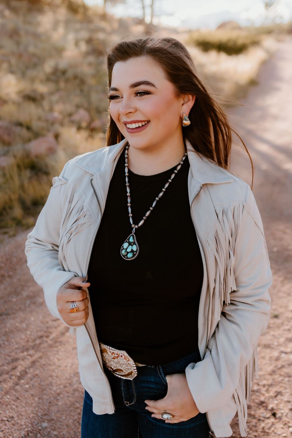 Smiling young woman in fringe jacket and turquoise necklace on a dirt road.