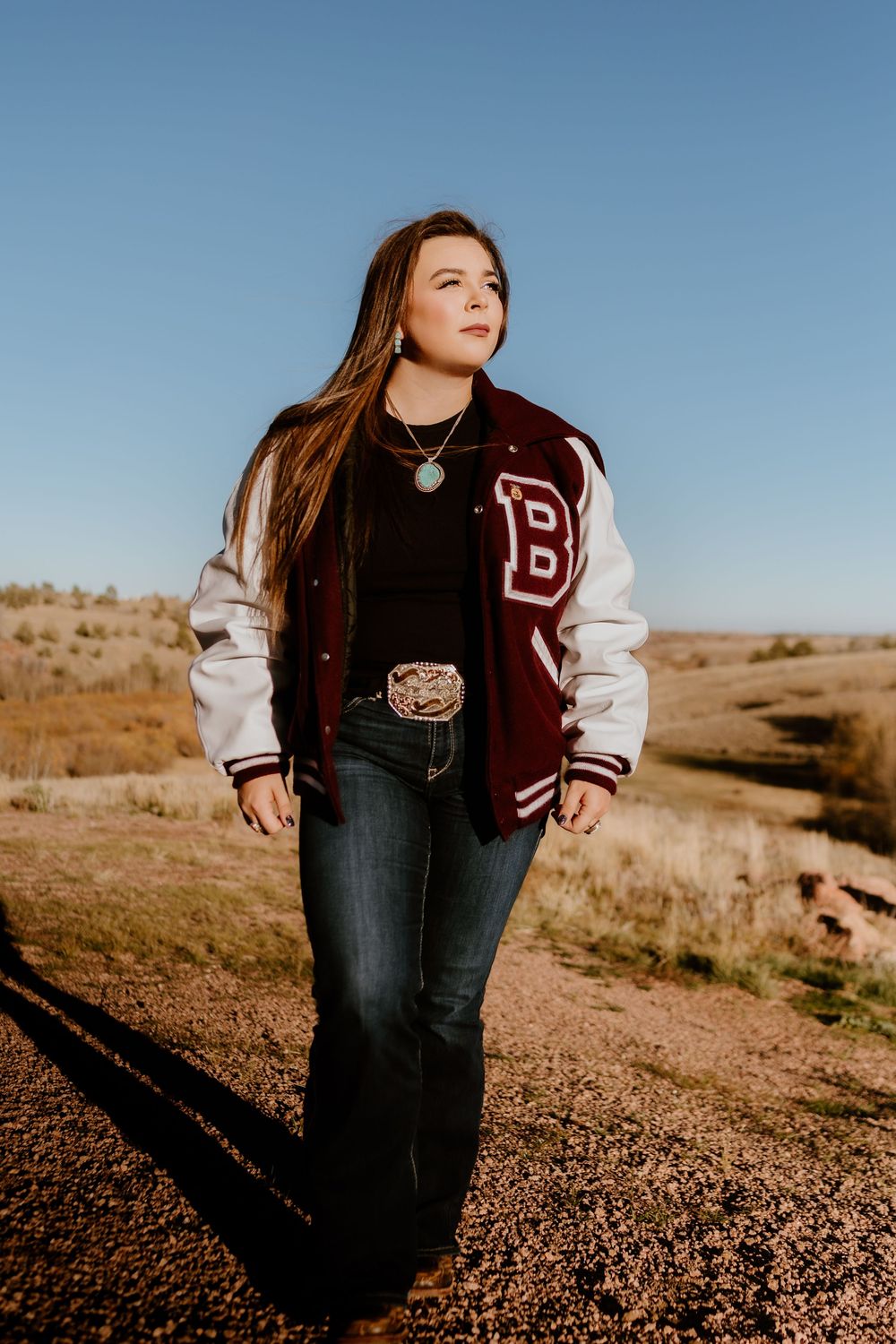 Young woman in varsity jacket with letter B, jeans, and turquoise necklace standing on rural dirt road under blue sky.
