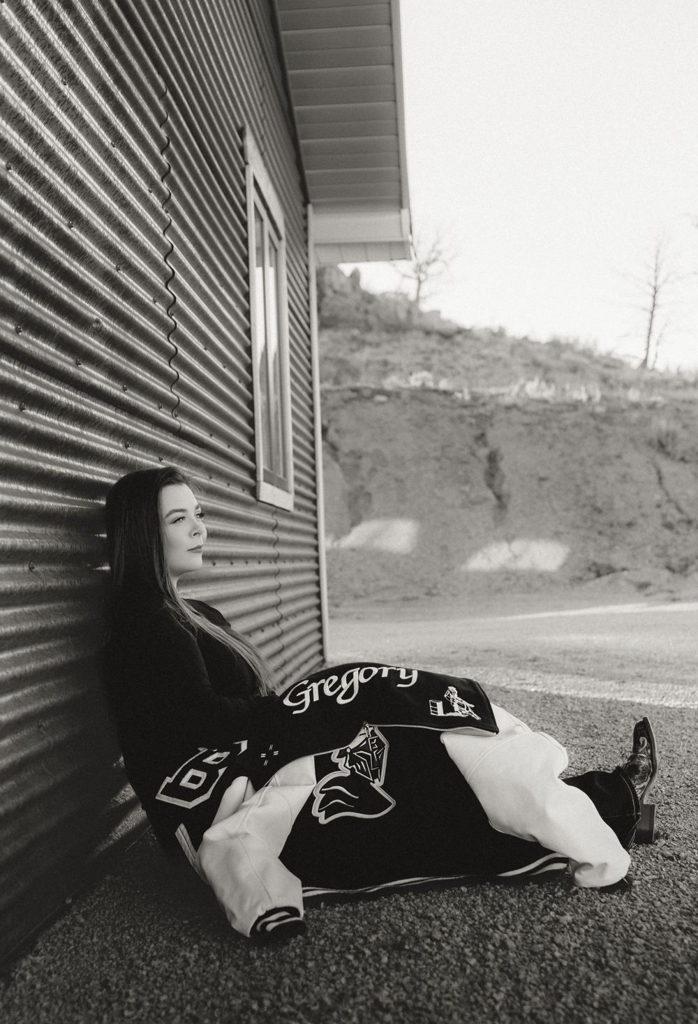 Young woman in letterman jacket sitting against corrugated metal wall in black and white photo.