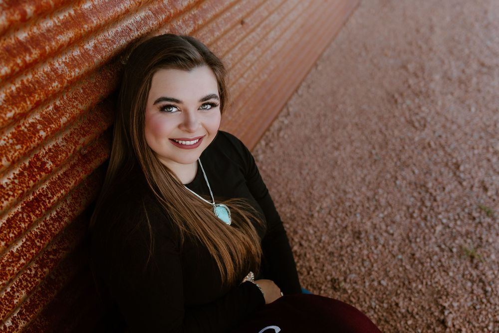Young woman with long brown hair smiles warmly, leaning against a rustic red wall, wearing a turquoise necklace.