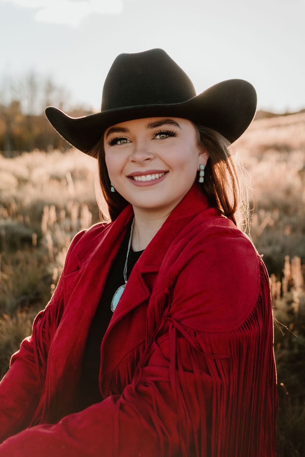 Smiling woman in red fringe jacket and black cowboy hat in a field at sunset.