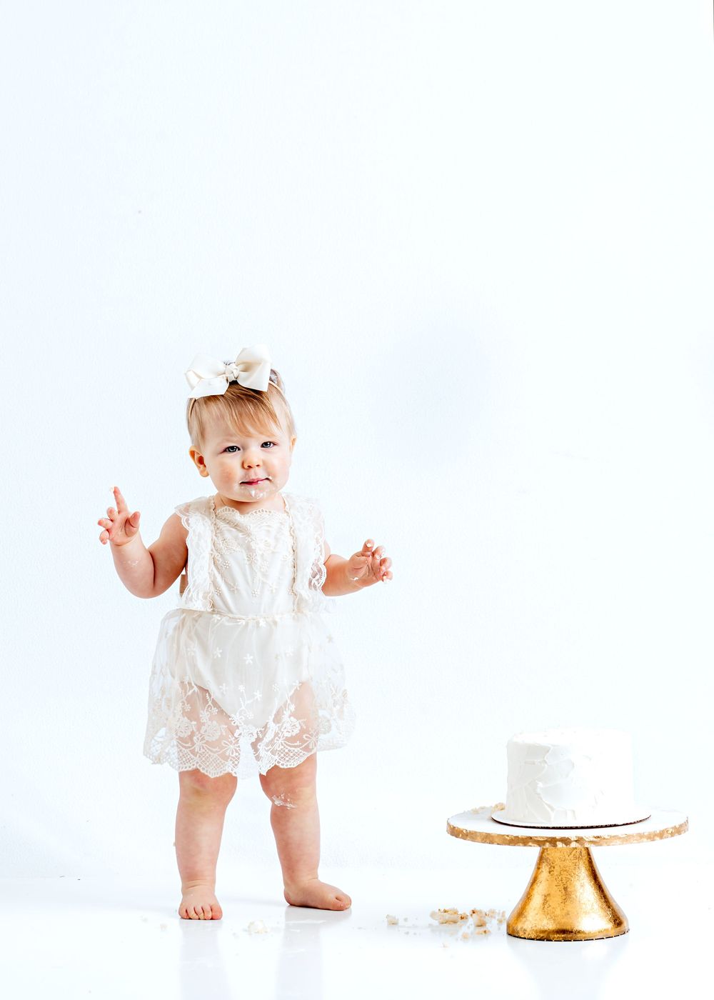A baby in a white lace dress stands next to a simple white cake on a gold cake stand against a white backdrop.