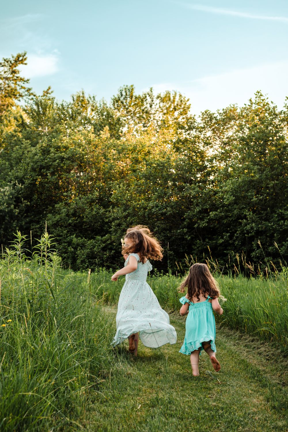 Two children in summer dresses run and dance through a sunlit grassy field bordered by green trees at sunset.