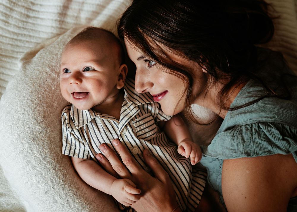 A smiling baby wearing a striped outfit lies on white bedding while being held lovingly from the side.