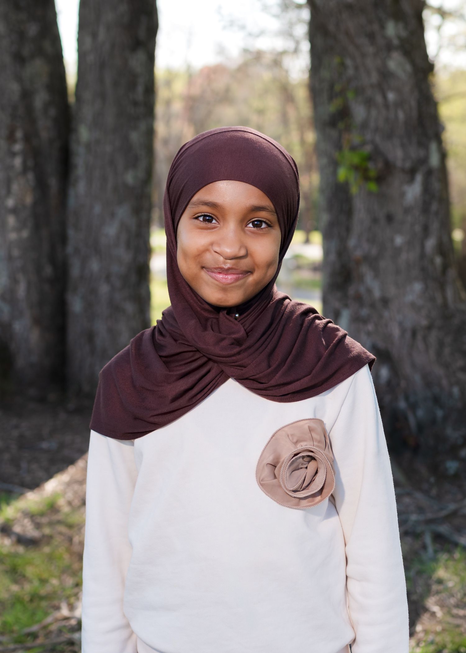Young girl wearing a brown hijab and white floral top smiling outdoors near trees.