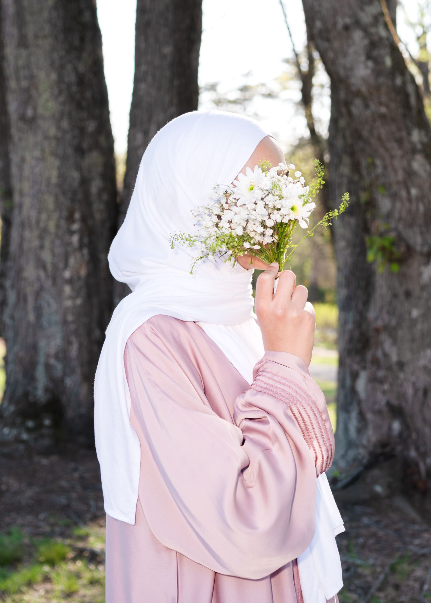 Woman in white hijab and pink abaya smelling white daisy bouquet outdoors.