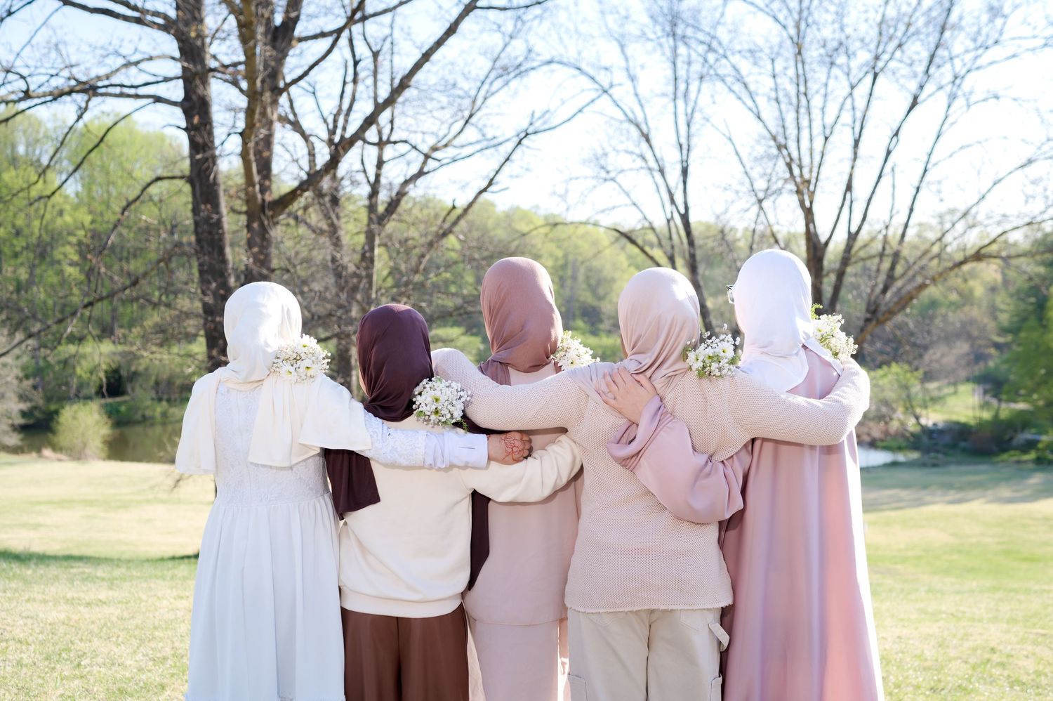 Five women wearing hijabs stand arm in arm outdoors, facing away from camera in a peaceful green park setting.