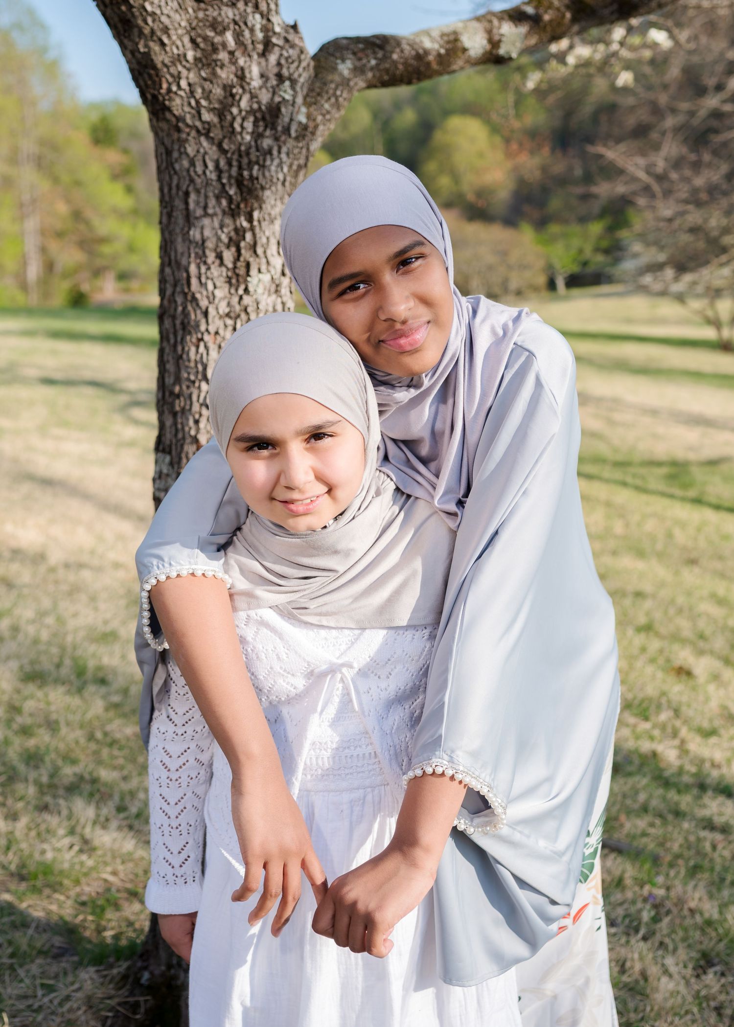 Two girls in matching gray hijabs embracing near a tree in a sunny park.