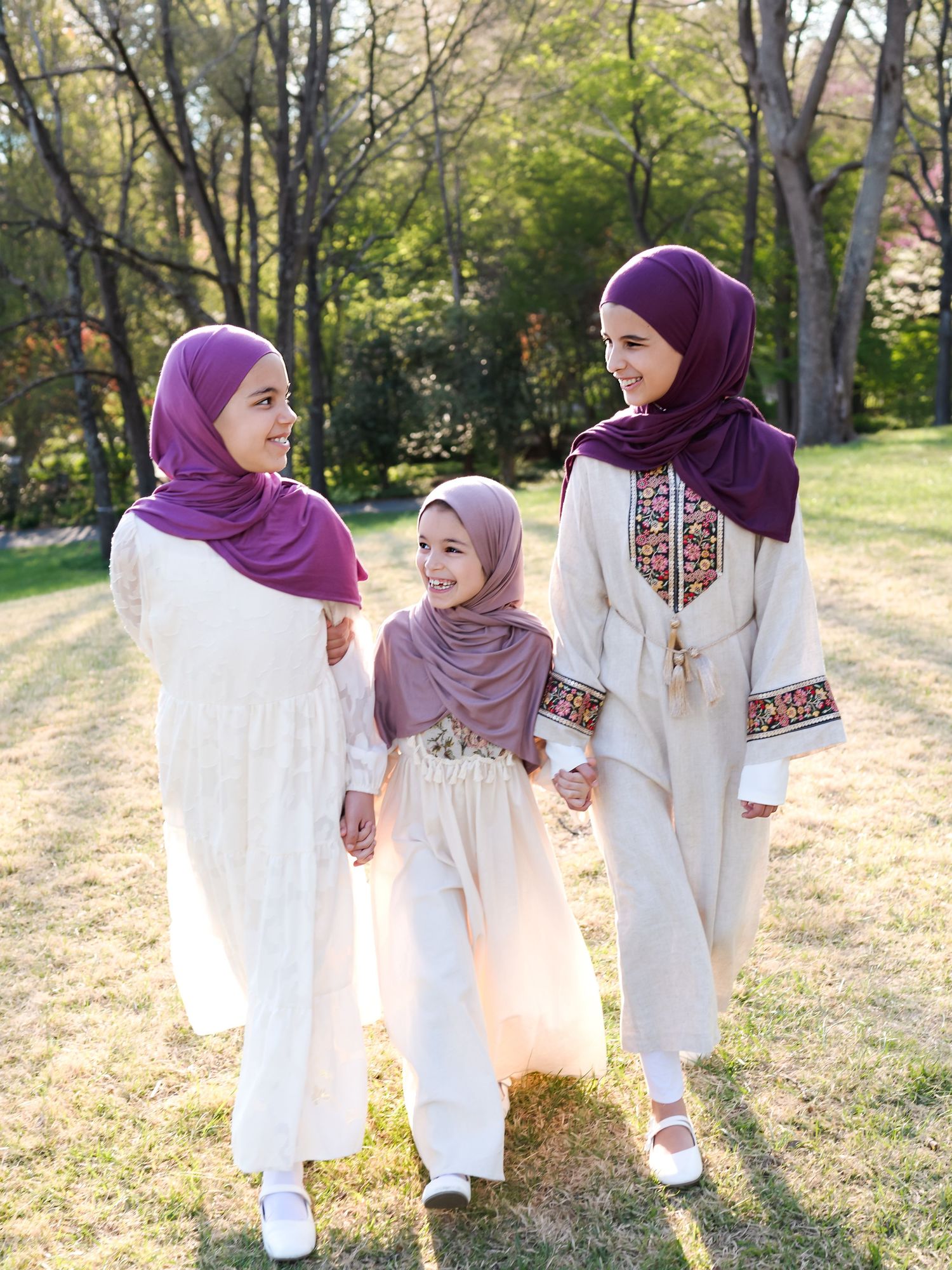 Three girls in matching cream outfits and purple hijabs walk together outdoors in a sunny park setting.