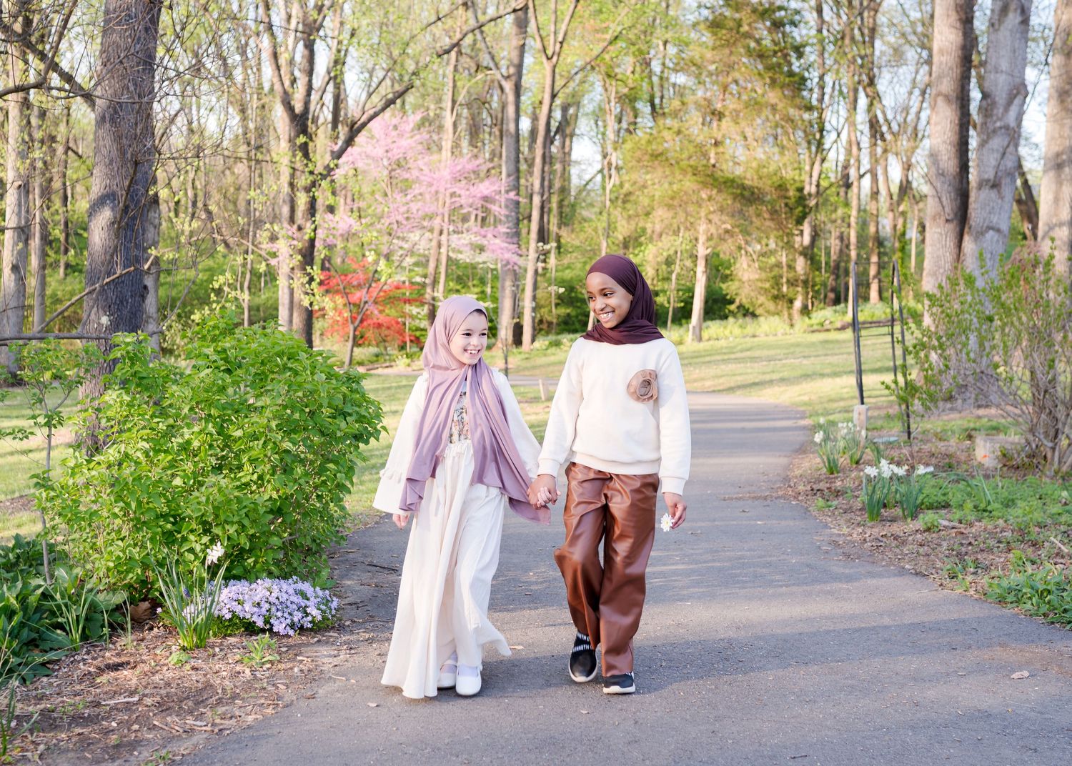 Two girls in hijabs walking hand in hand on a sunny park path.