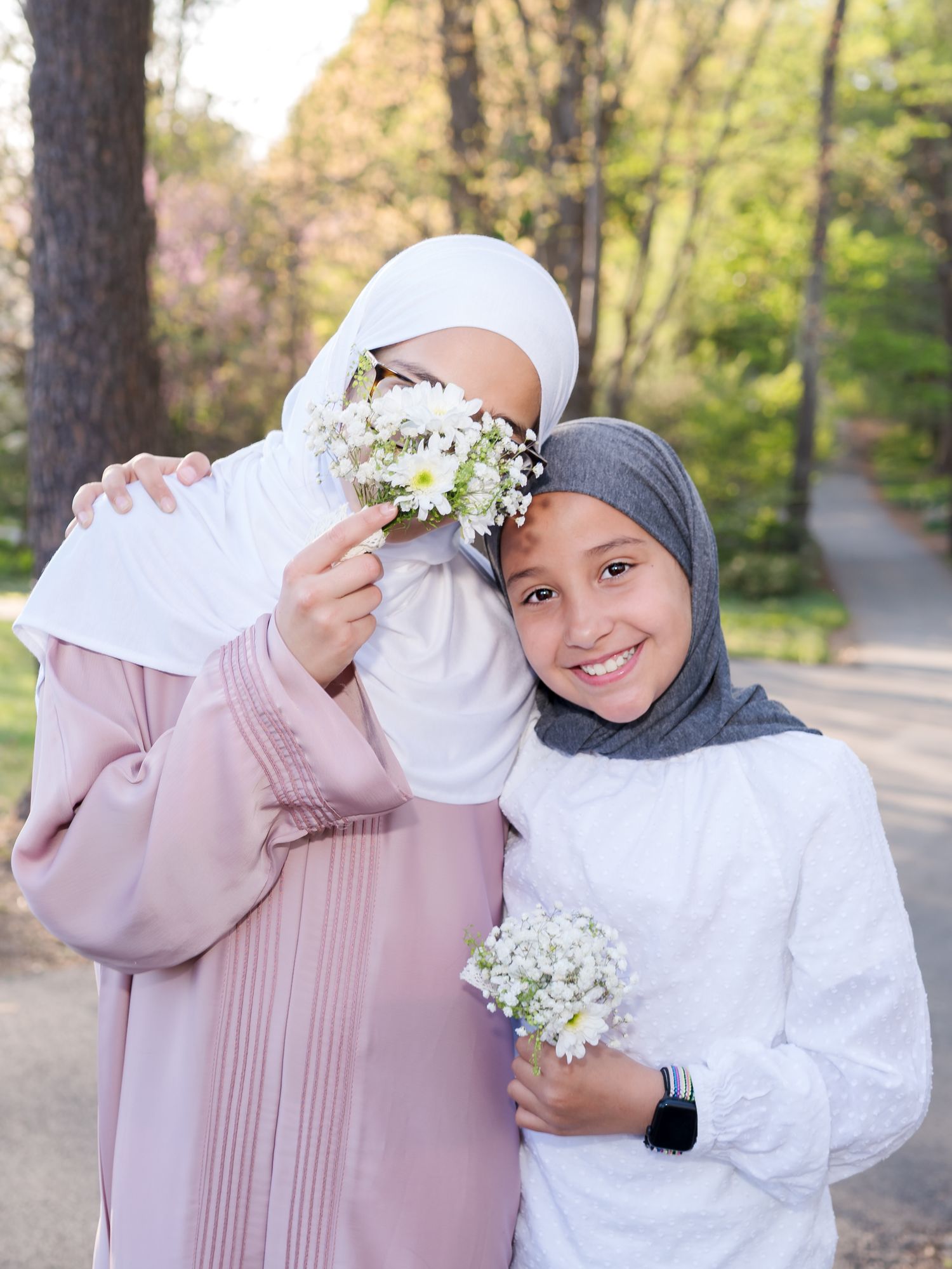 Two girls in hijabs holding white flowers, smiling in a park setting with trees in the background.