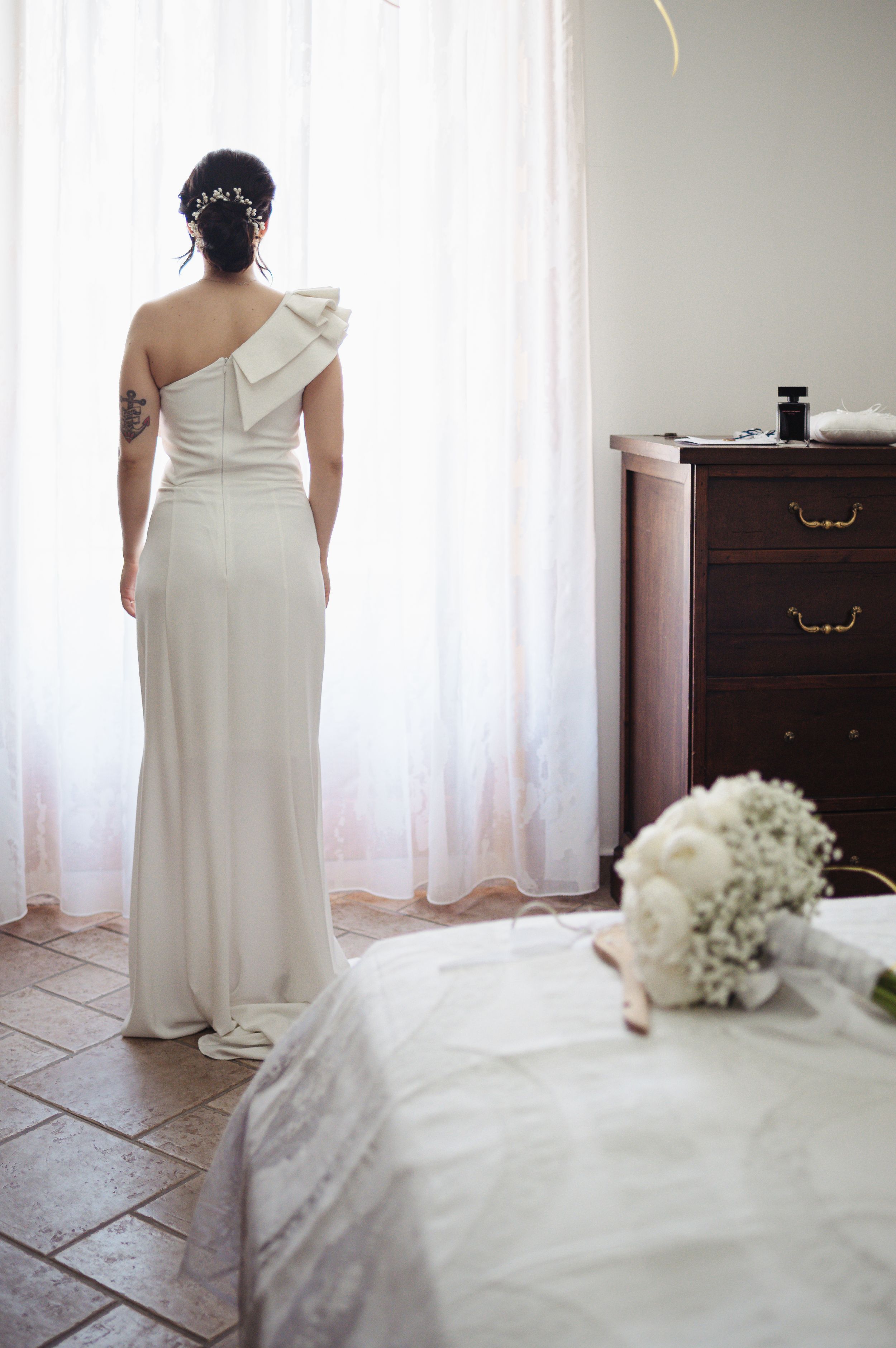 Back view of elegant white wedding gown with asymmetrical neckline and white floral bouquet visible in foreground.