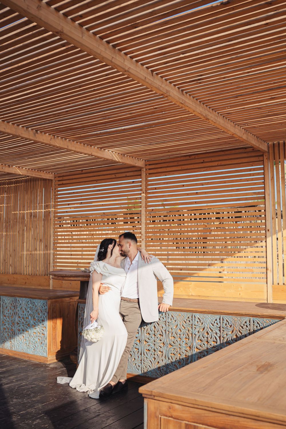 Couple shares romantic moment on wooden deck with slatted ceiling and geometric patterned wall.
