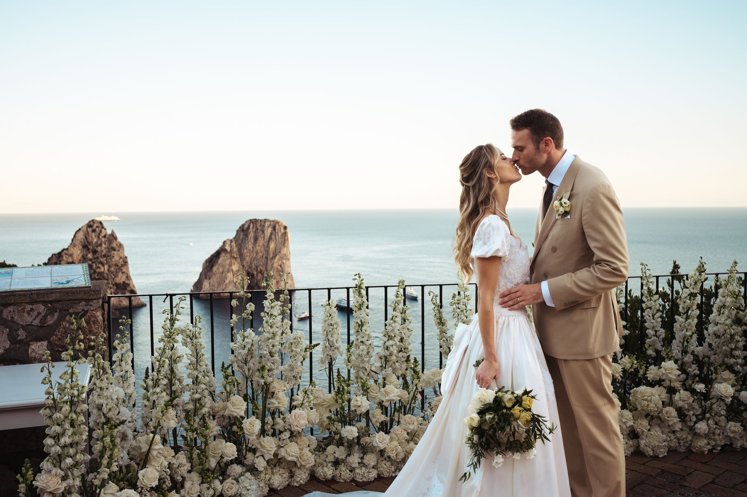 Romantic wedding couple embraces by ocean overlook with stone wall and sunset backdrop.