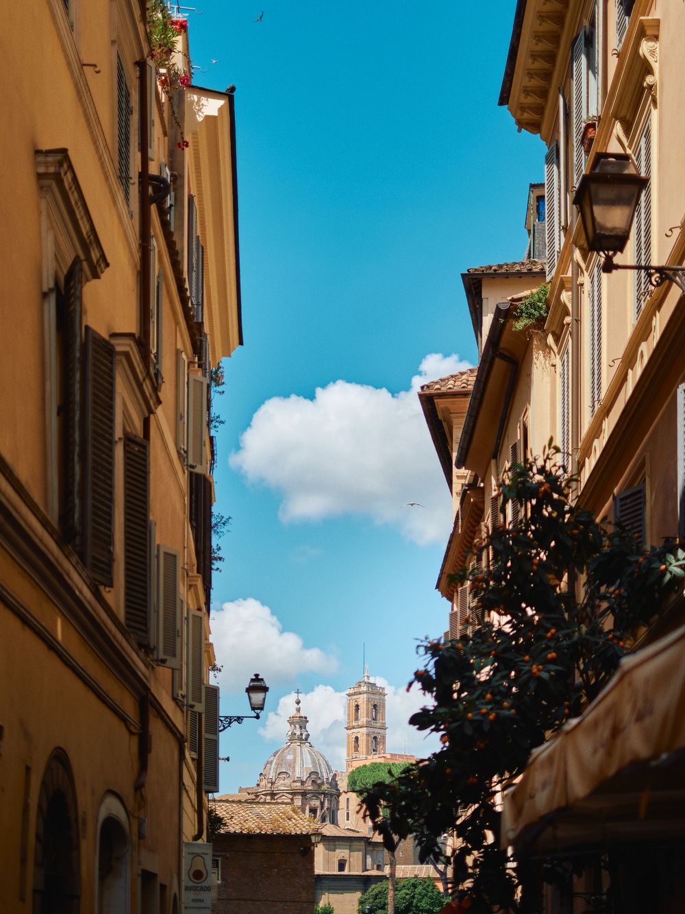 Narrow Italian street in Rome with yellow buildings and church bell tower visible against bright blue sky with clouds.