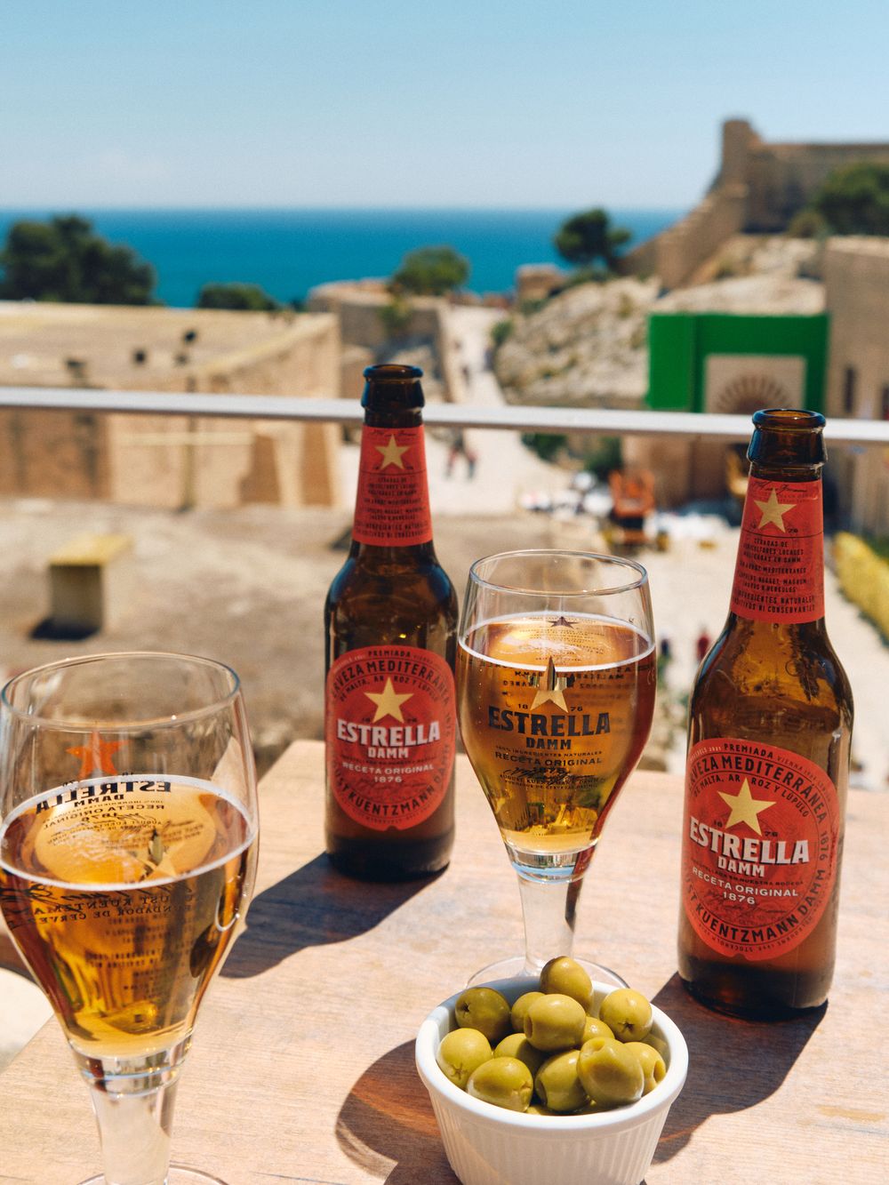 Cold bottles of Estrella beer with green olives and views of Mediterranean coastline and castle on sunny terrace.