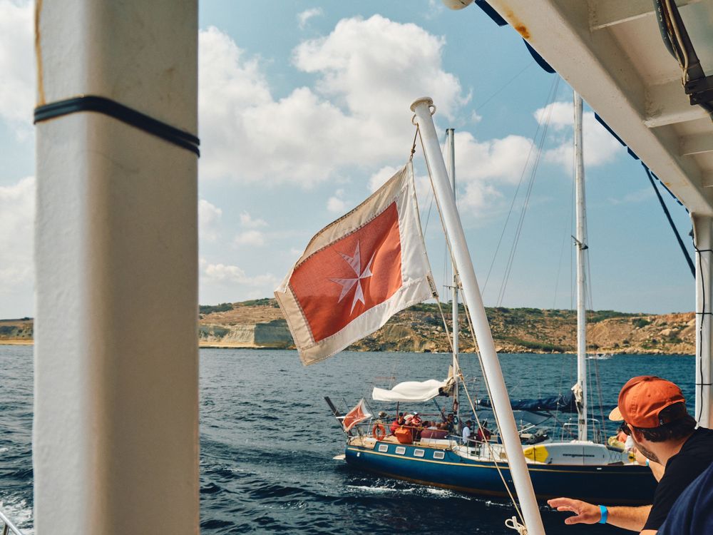 A red flag waves from a sailboat on blue waters while another boat with orange-clad passengers cruises nearby.