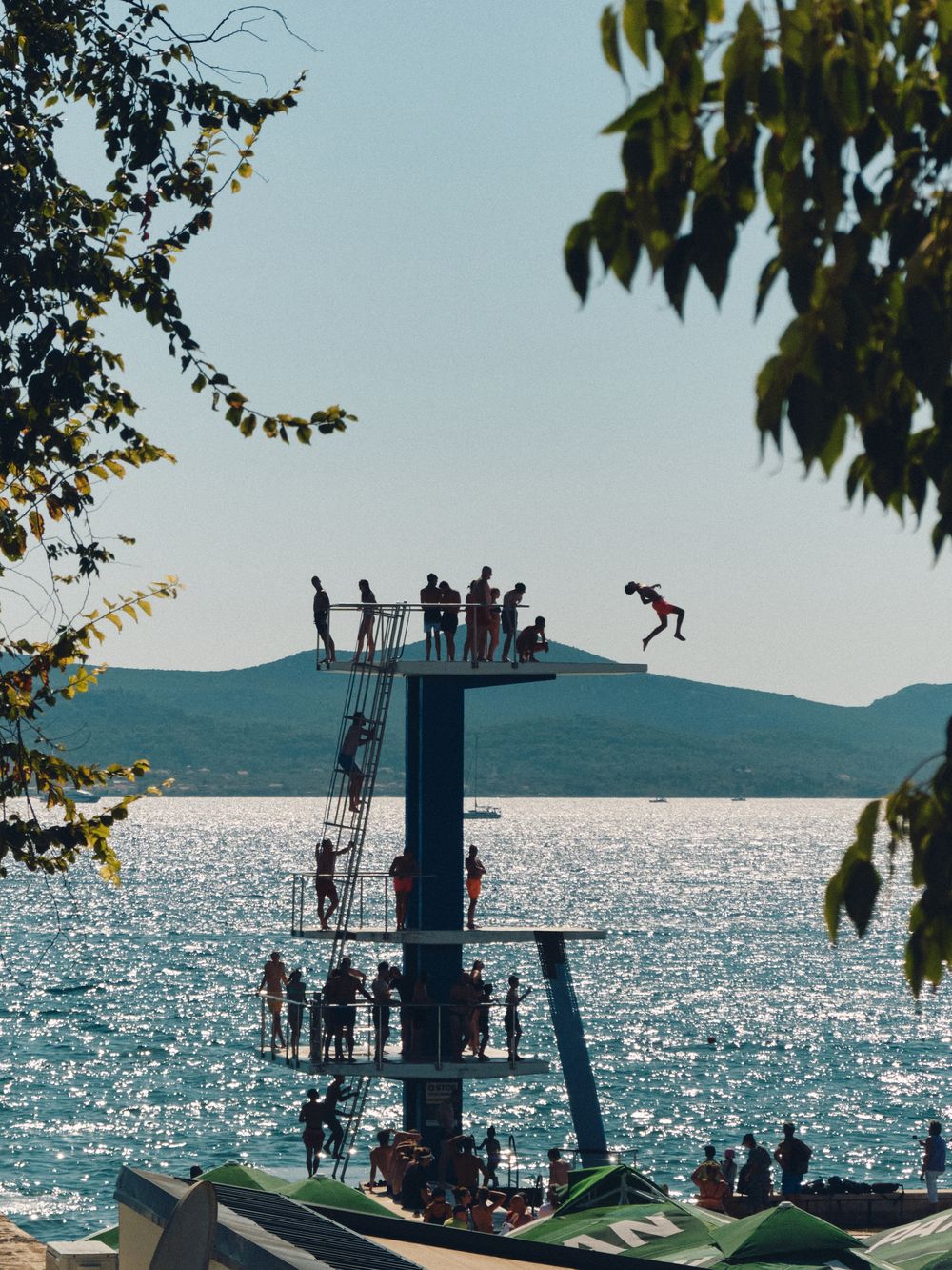 People silhouetted on diving platforms against sparkling ocean waters and mountain backdrop at sunset.