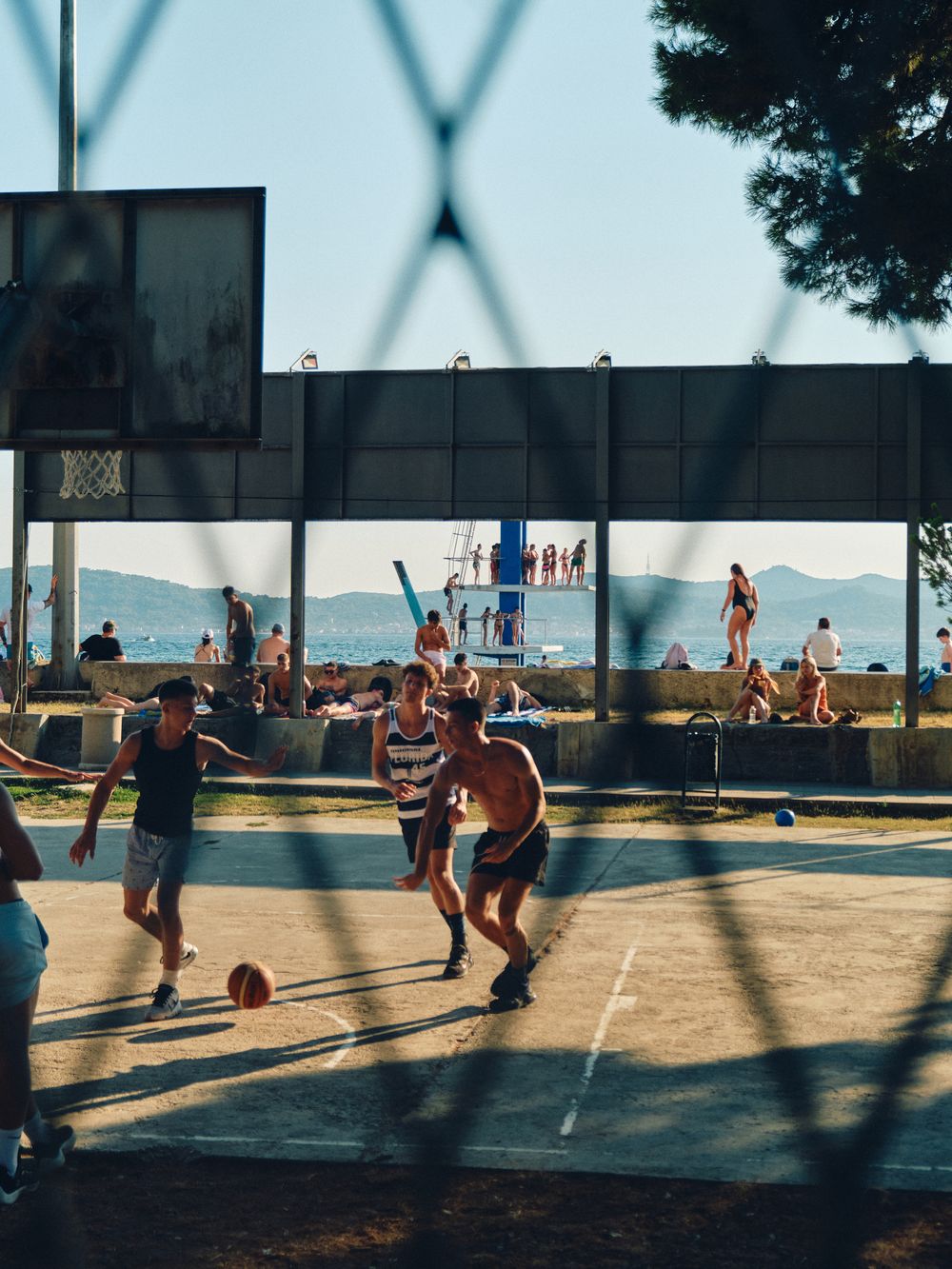 People playing volleyball at a beachside court during sunset casting long shadows across the sandy surface.