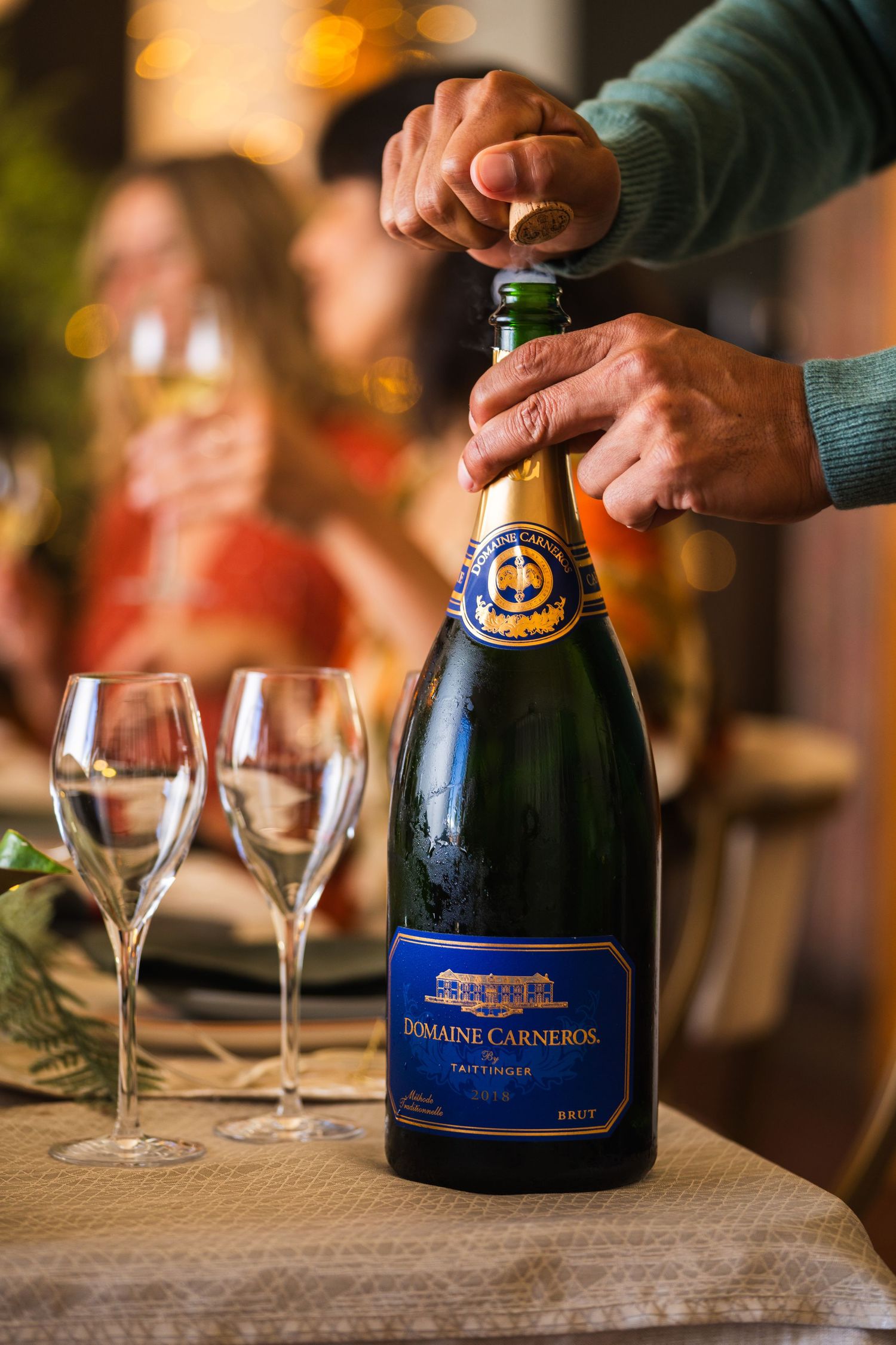 Hands opening a bottle of Domain Carneros sparkling wine with empty champagne flutes on wooden table.