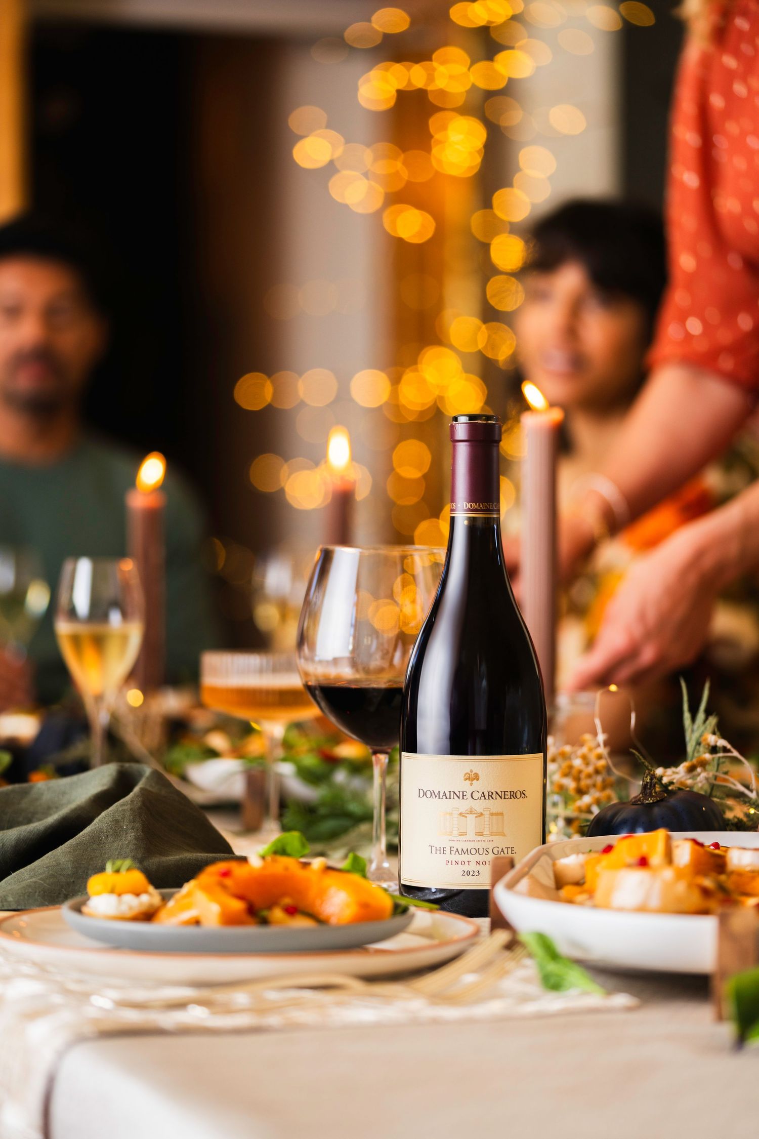 Wine bottle and elegant table setting at dinner party with glowing candles and festive bokeh lights in background.