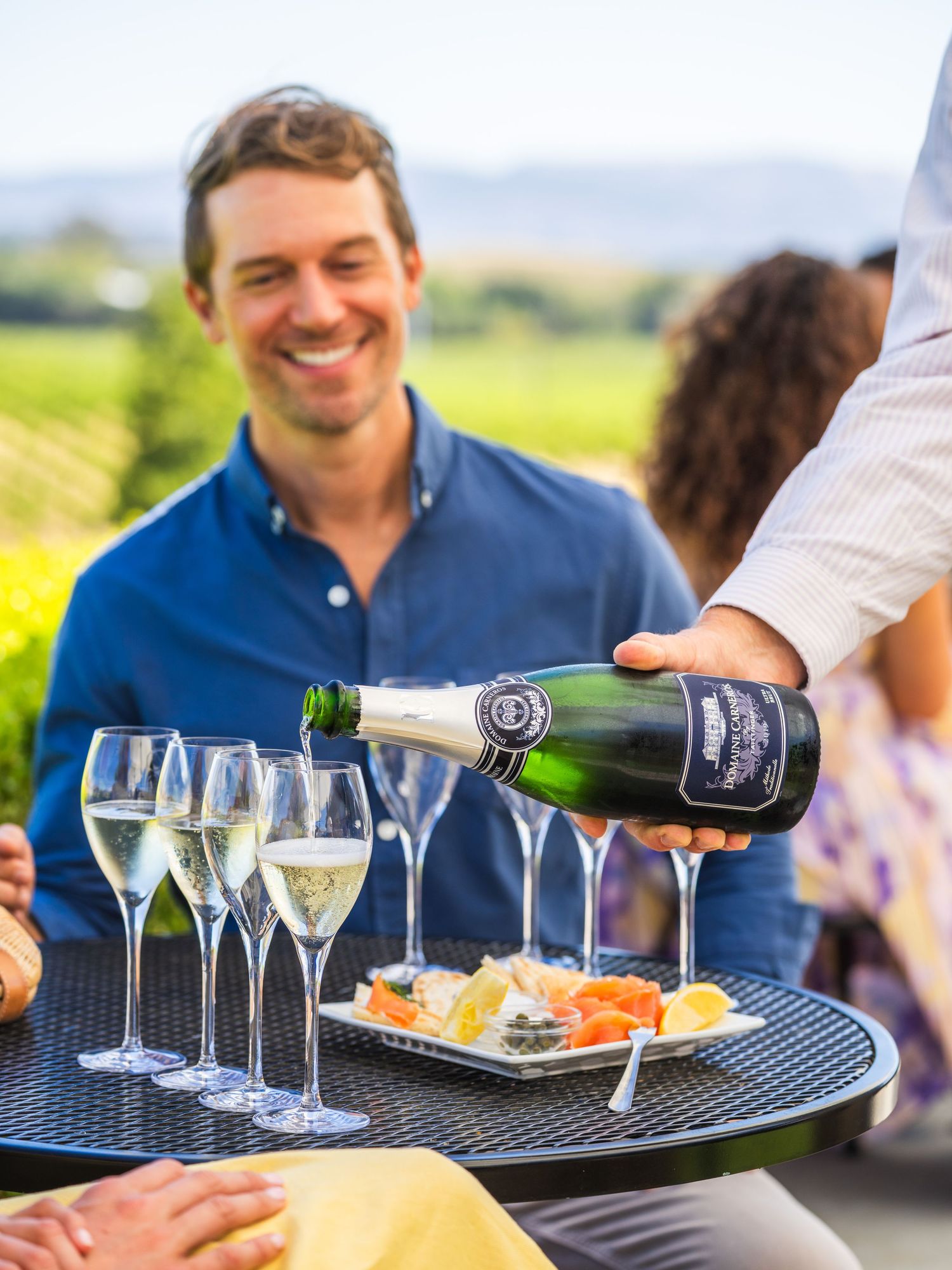 A waiter pours champagne into glasses at an outdoor party while guests enjoy appetizers on a sunny day.