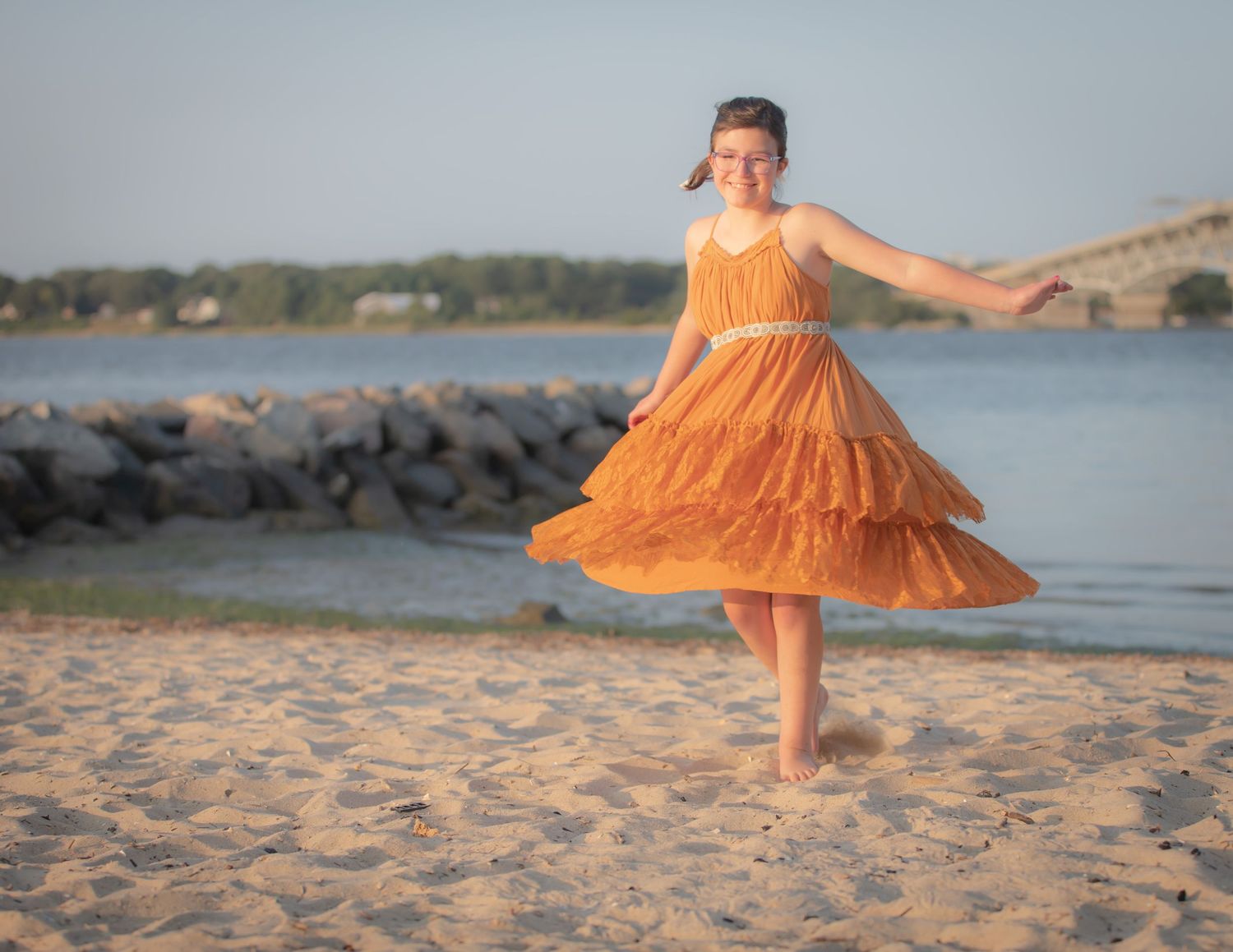 Young girl in an orange tiered dress twirling joyfully on a sandy beach near the water at sunset.