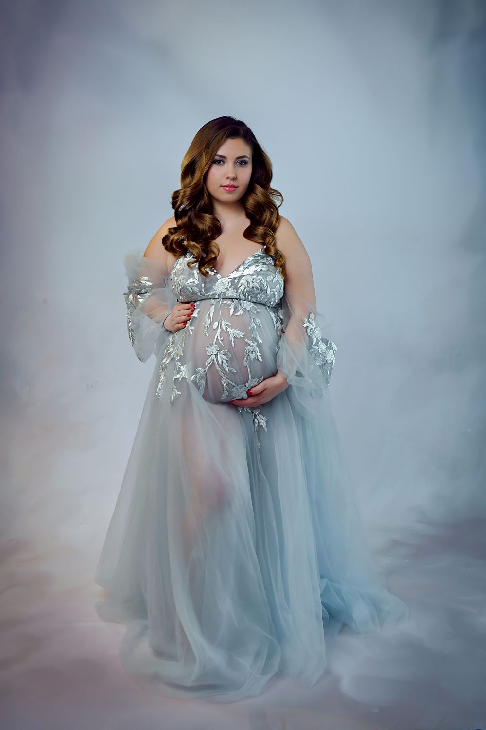 Ethereal maternity photo featuring a flowing blue-gray tulle gown with lace details in a dreamy studio setting.