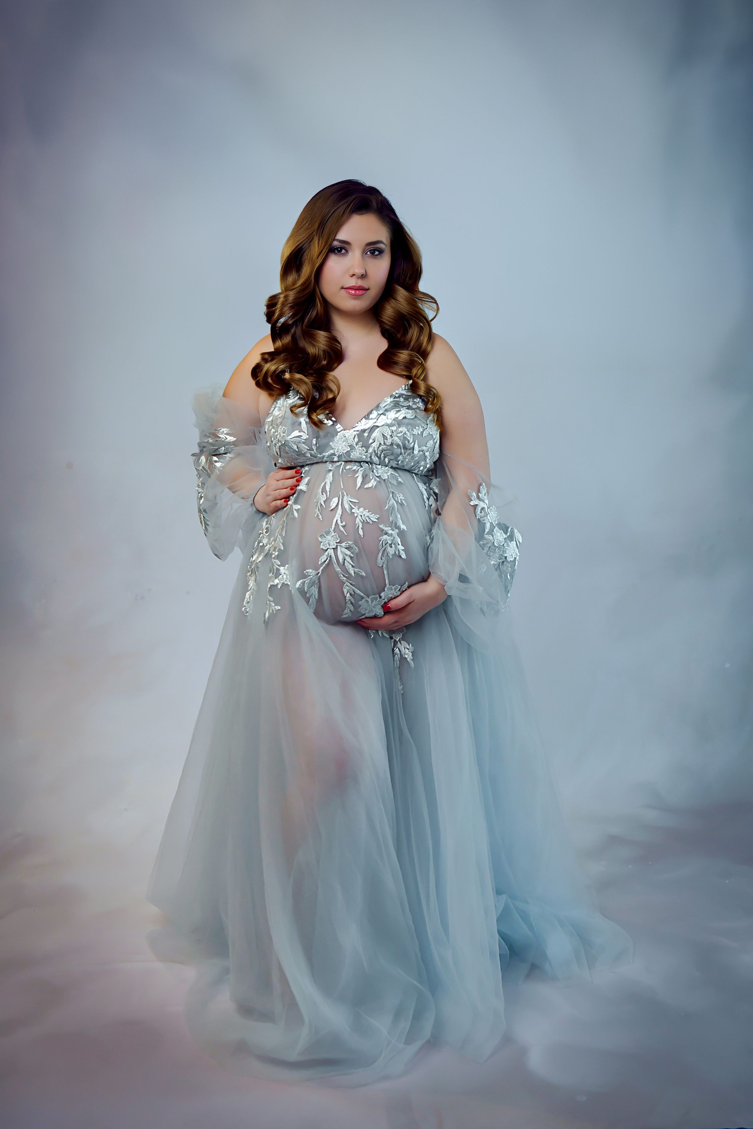 Ethereal maternity photo featuring a flowing blue-gray tulle gown with lace details in a dreamy studio setting.