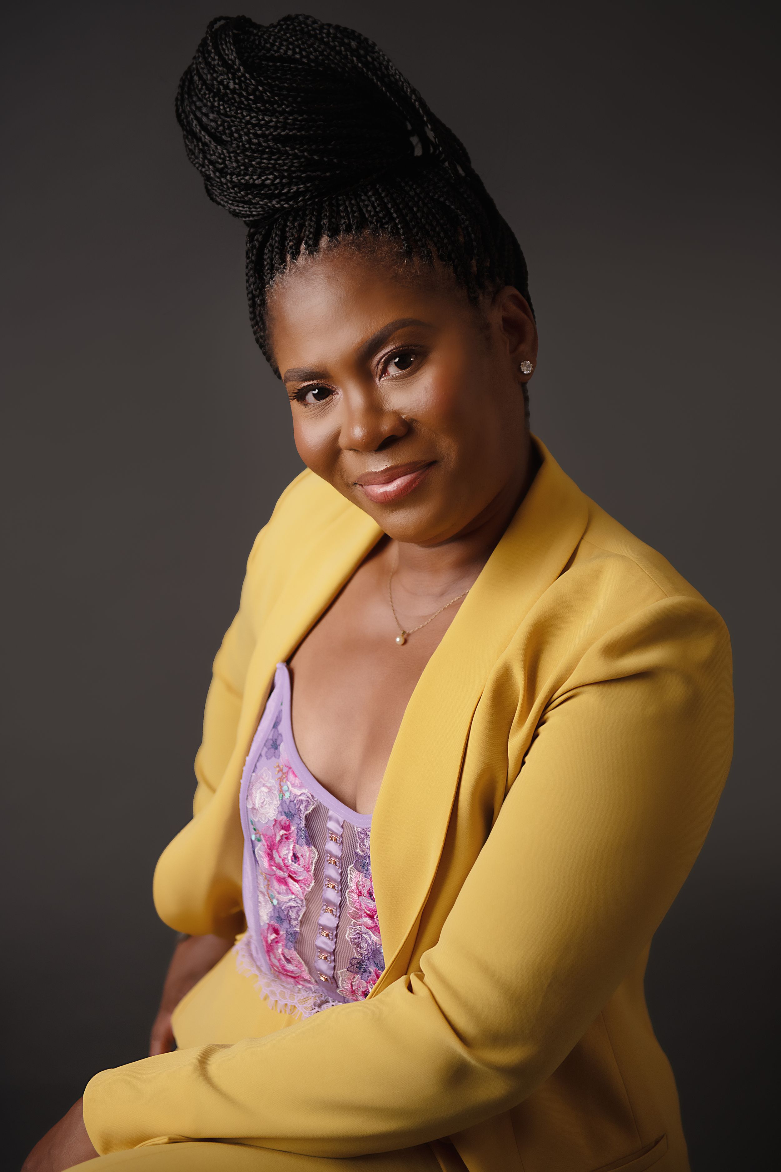 Confident woman in yellow blazer and braided updo poses against dark backdrop.