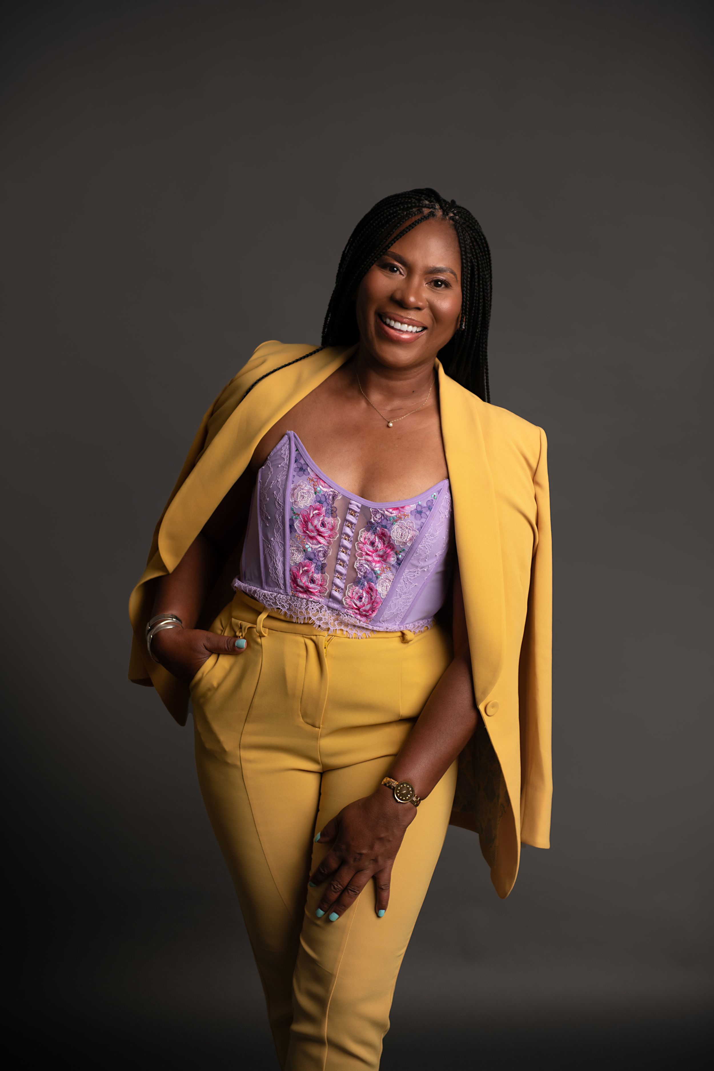 Professional portrait of model in yellow blazer and pants with purple corset top against dark background.