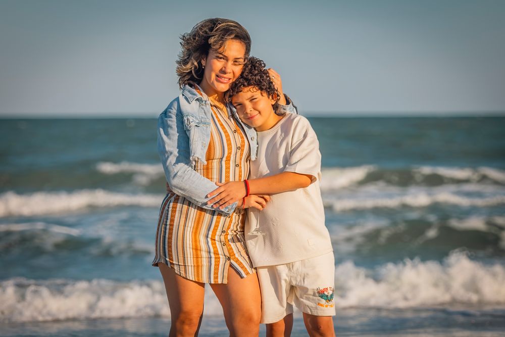 A woman and child embrace warmly on a sunny beach, waves crashing behind them, wearing casual summer outfits.