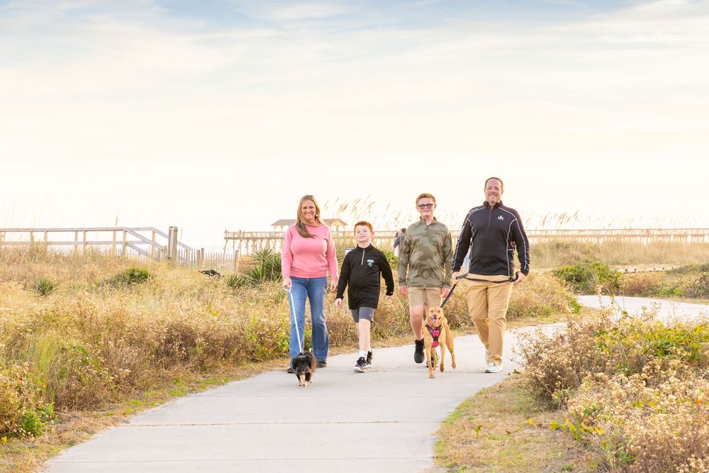 Family of three with a dog walking on a coastal path surrounded by dunes and greenery on a sunny day.