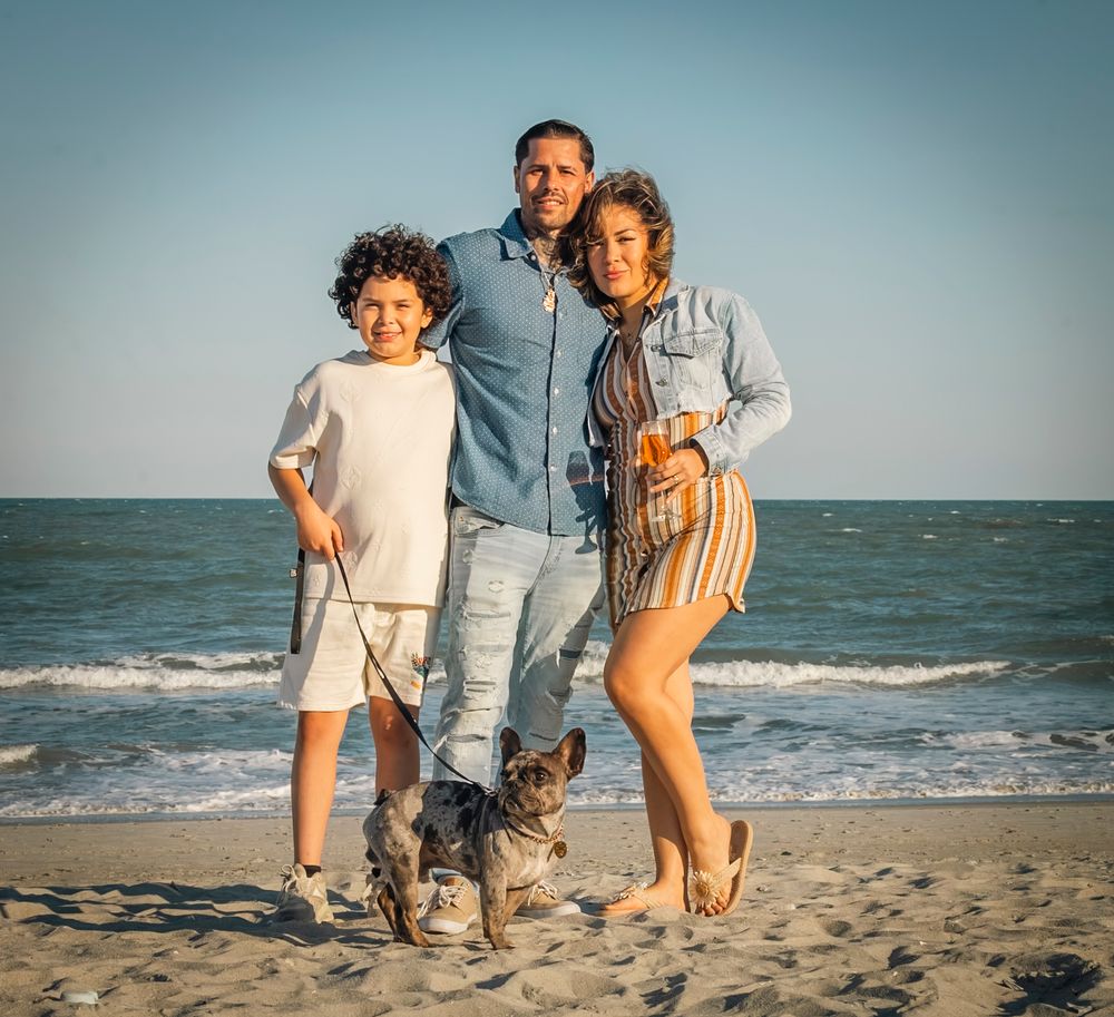 Family of three with a dog posing on a sandy beach with ocean waves in the background on a sunny day.