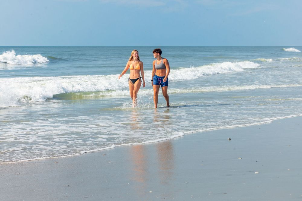 Couple walking along sandy beach shoreline in swimwear with ocean waves in background on sunny day.