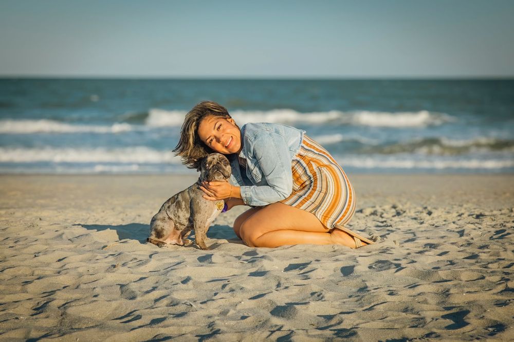 Woman in denim shirt and striped skirt smiling while petting a small dog on a sunny sandy beach.
