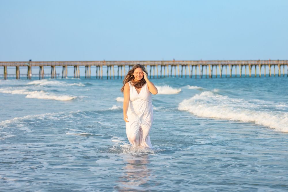A woman in a white dress runs joyfully through ocean waves near a pier on a sunny beach day.