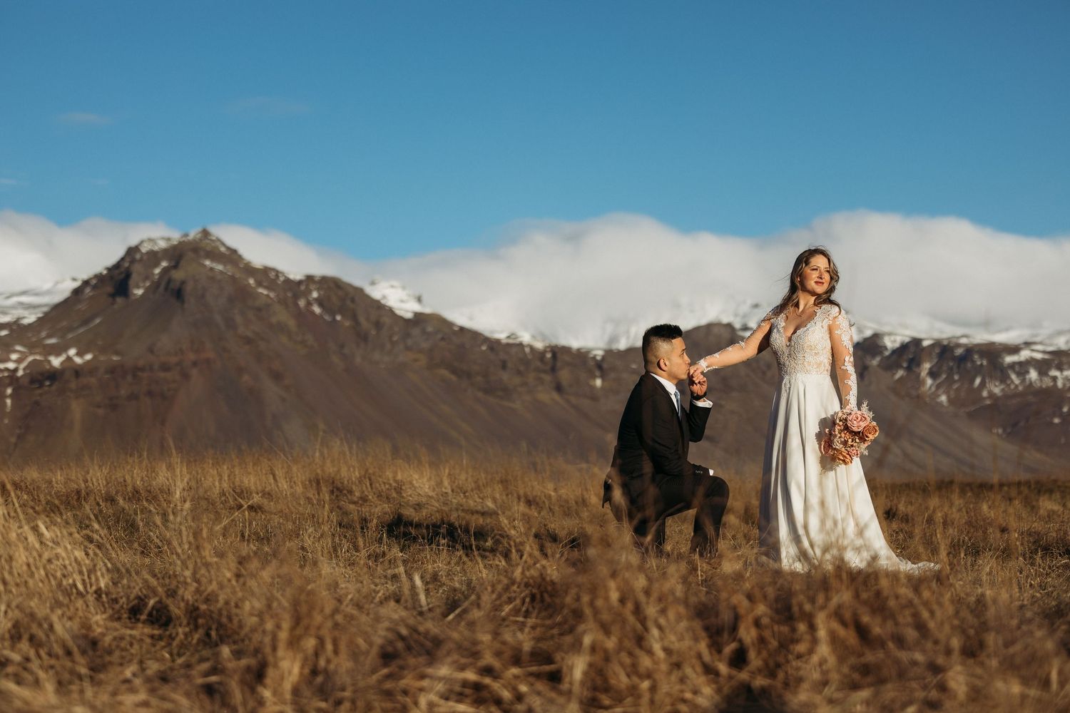 Couple in wedding attire posing in Iceland's dramatic landscape with snow-capped mountains and golden fields.