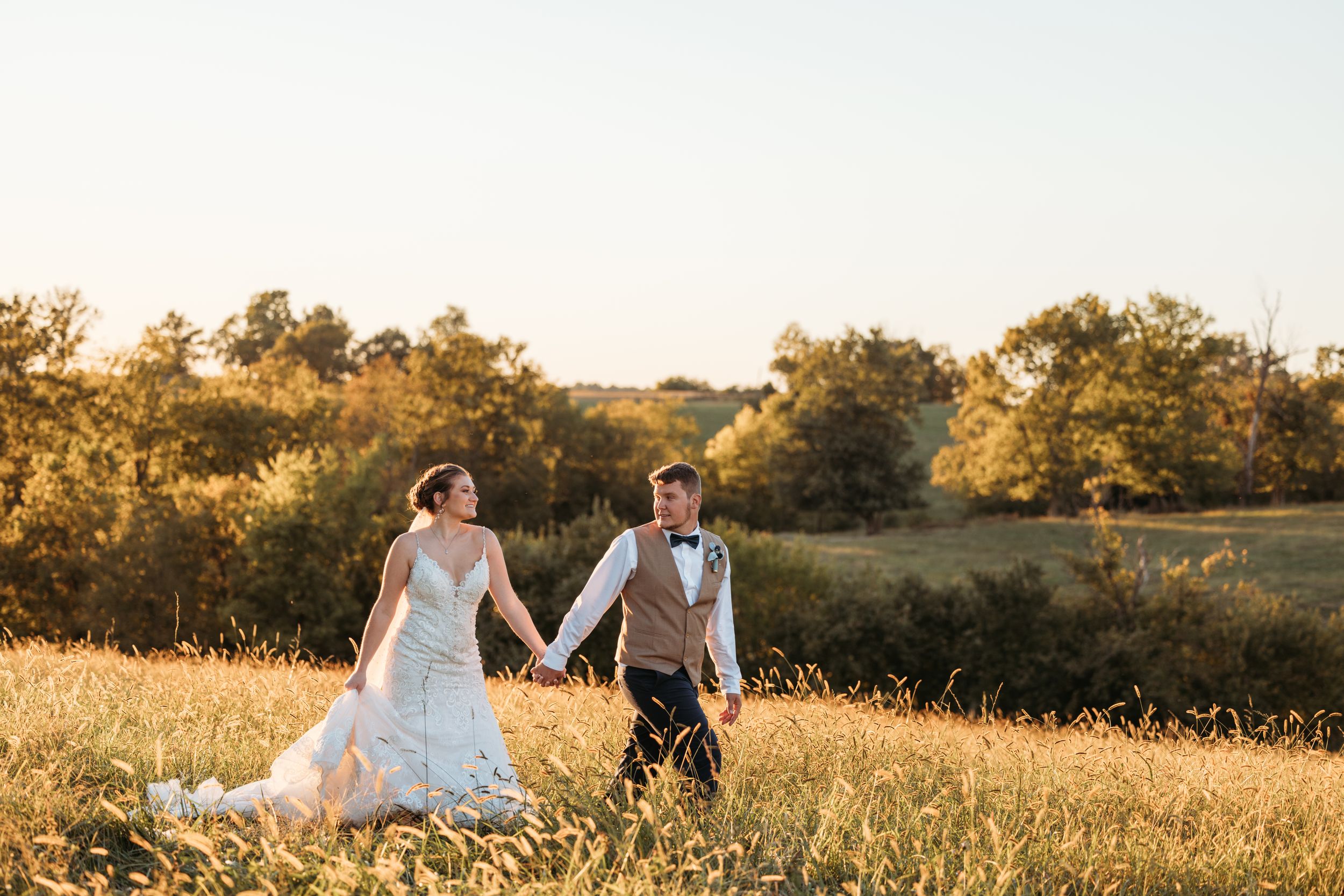 Bride and groom holding hands walking through golden sunlit field at sunset on their wedding day.