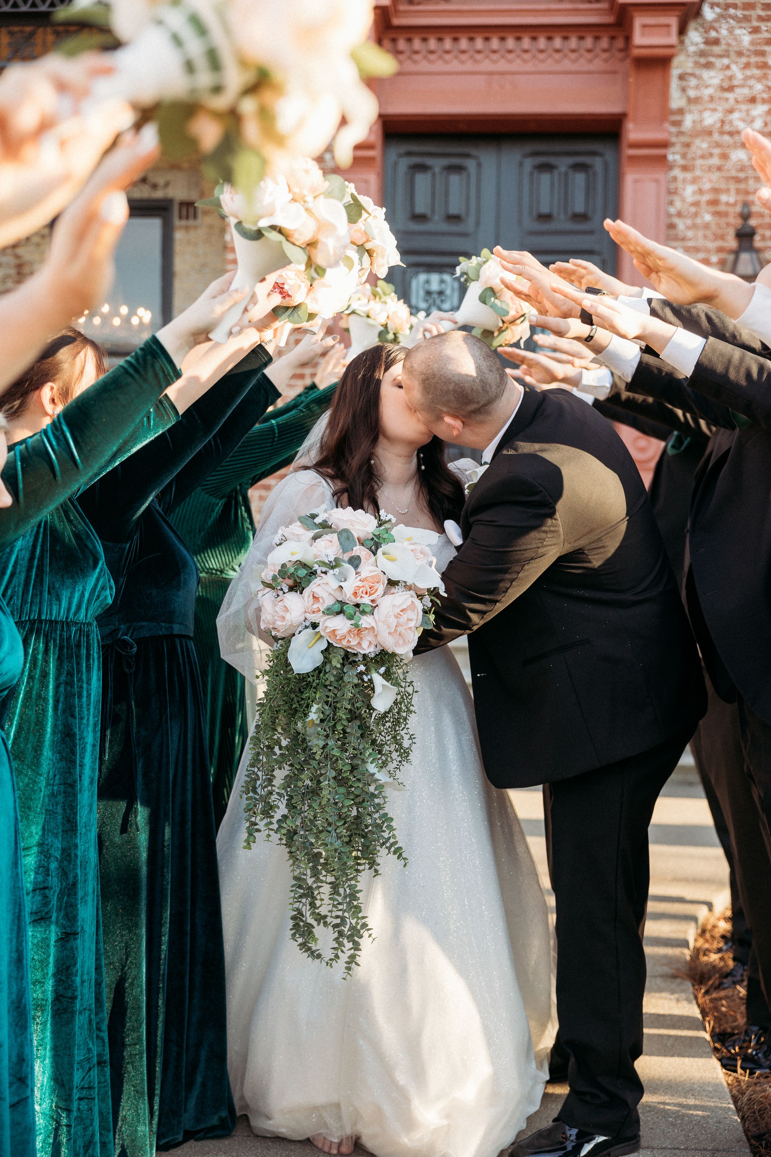 Bride and groom kiss under an arch of bouquets held by bridesmaids in emerald green velvet dresses outside a brick building.