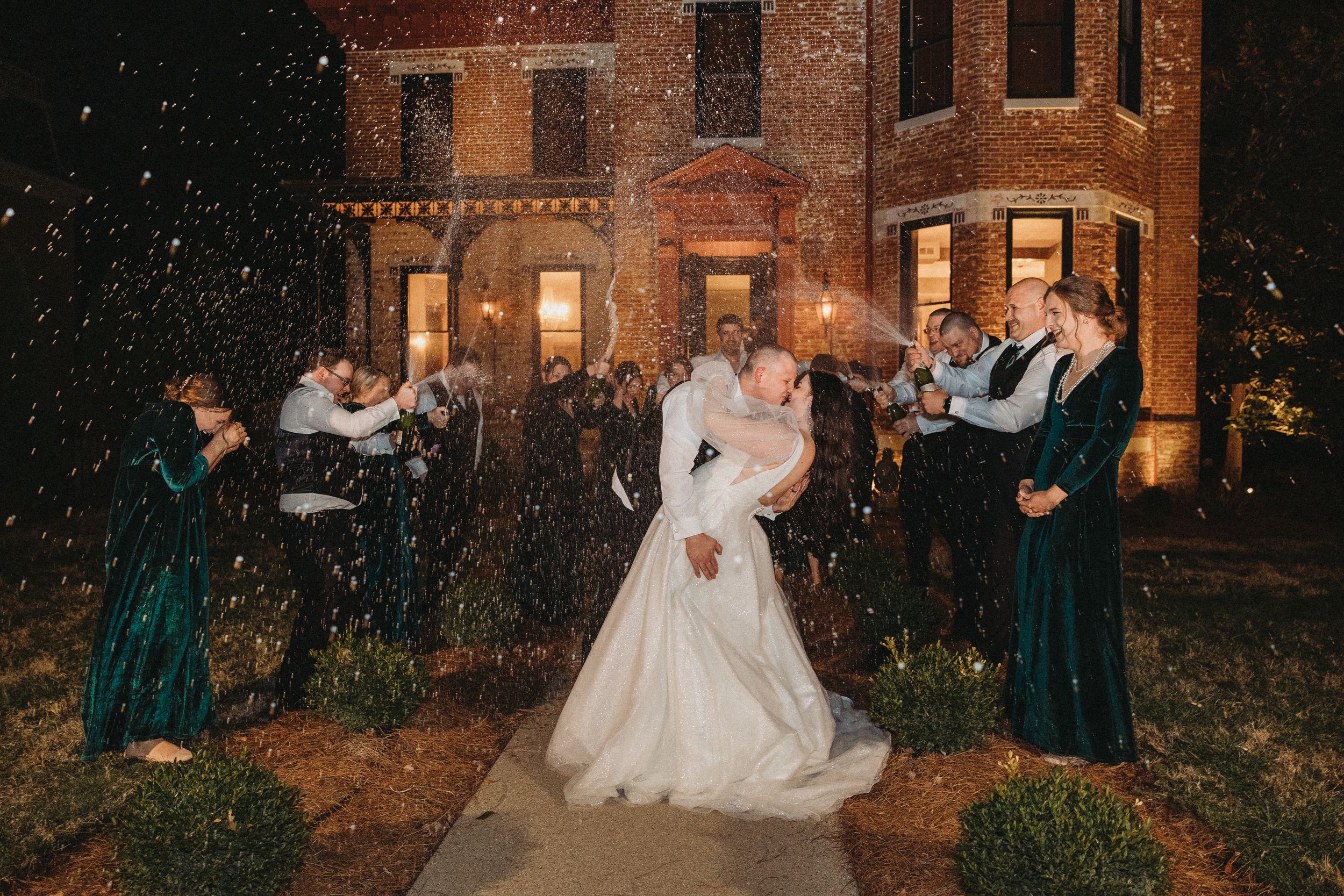 Bride and groom share a kiss while wedding party celebrates with champagne spray outside a brick mansion at night.