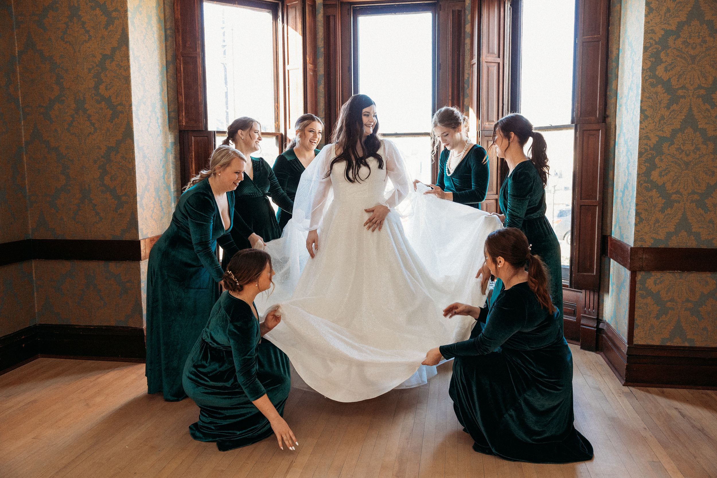 Bride in white gown surrounded by bridesmaids in emerald green velvet dresses arranging her skirt in elegant room.