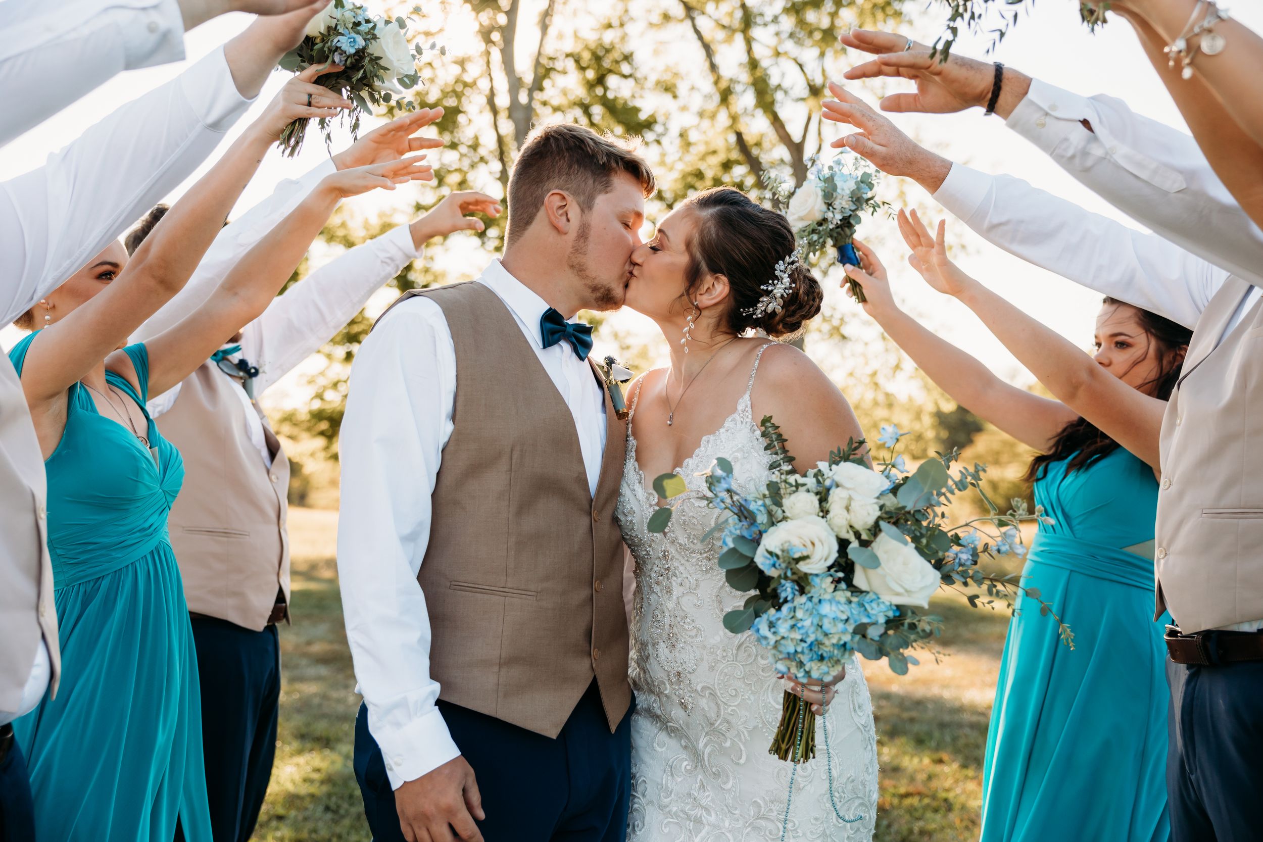 Bride and groom kissing under an arch of raised hands formed by wedding party in teal dresses and tan vests outdoors.