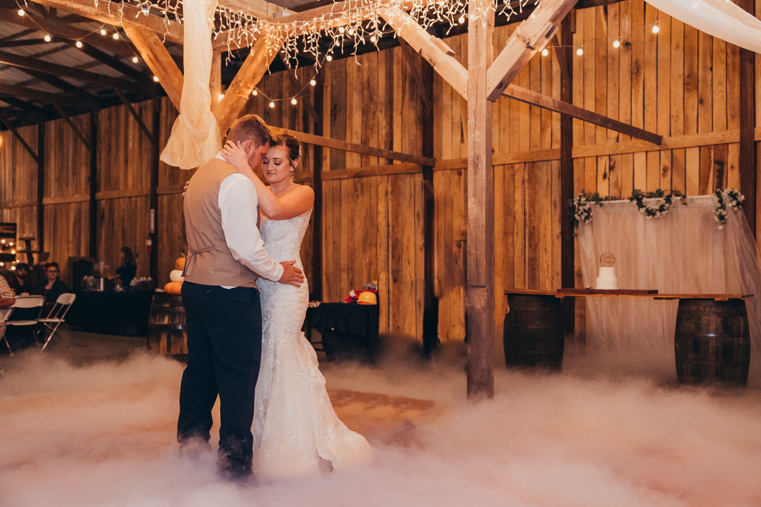 Bride and groom share first dance in rustic barn venue with fog machine and twinkling lights.