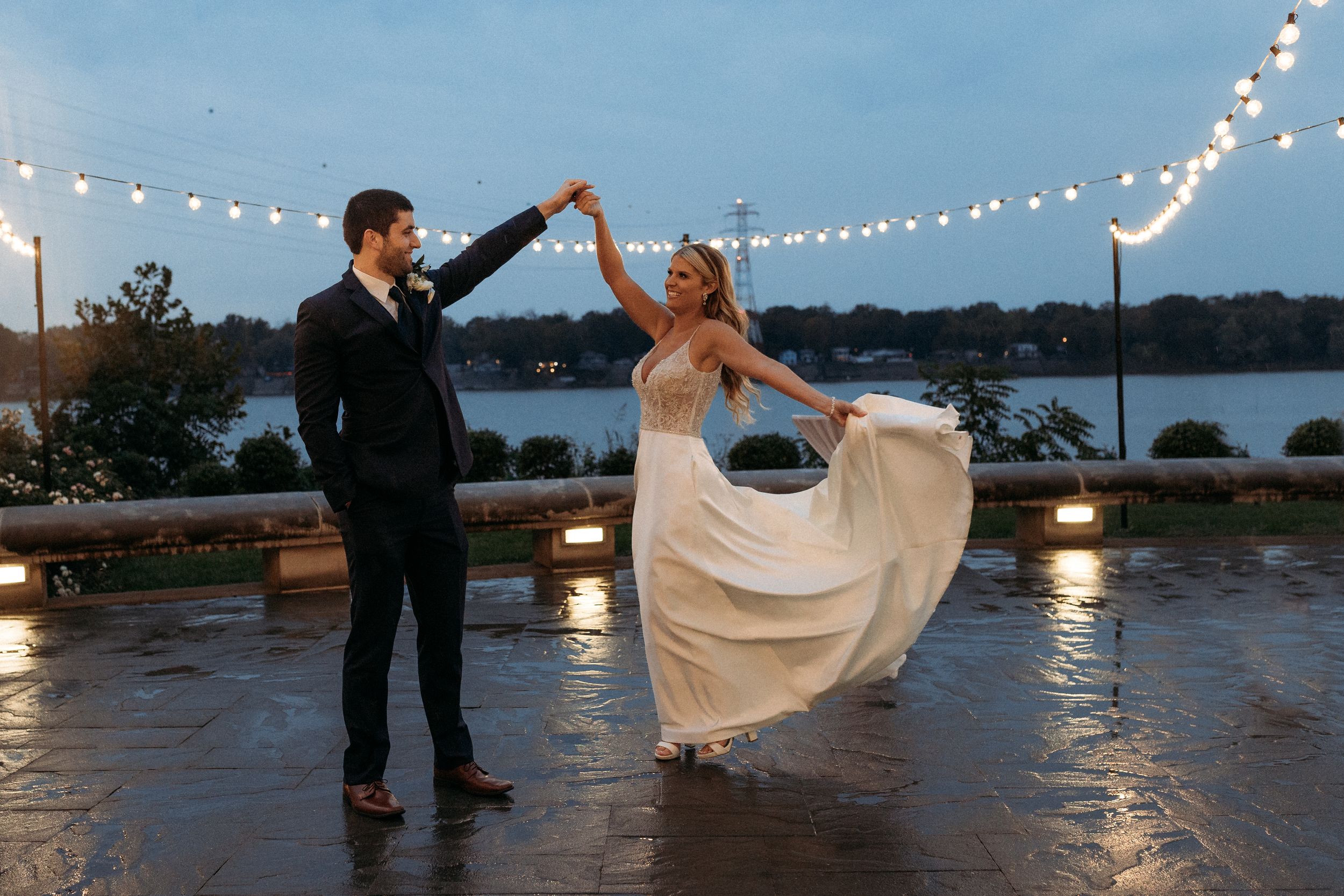 Bride and groom dancing outdoors at dusk near a waterfront, surrounded by string lights in wedding attire.