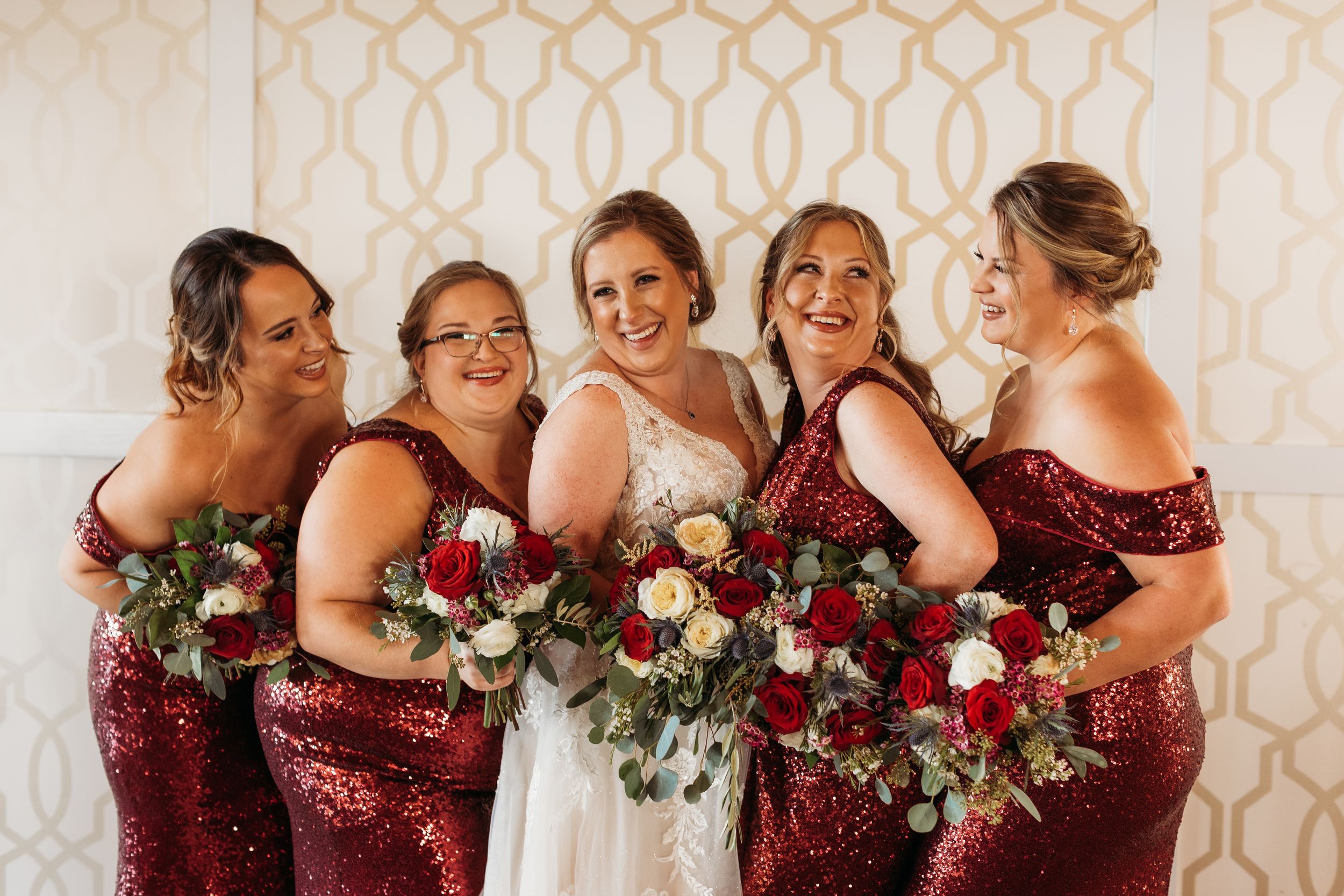 Bride in white gown with four bridesmaids in red sequin off-shoulder dresses holding bouquets of red and white roses.