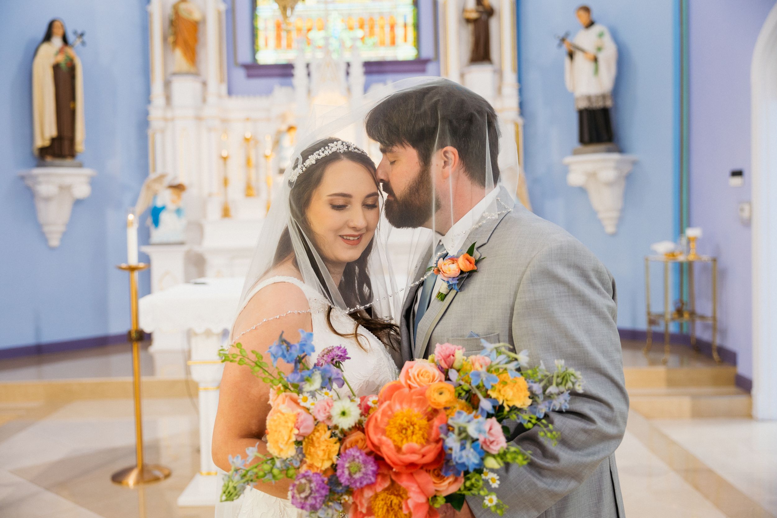 Bride and groom share a tender moment in a blue church, bride holding vibrant colorful bouquet of orange, pink, and blue flowers.