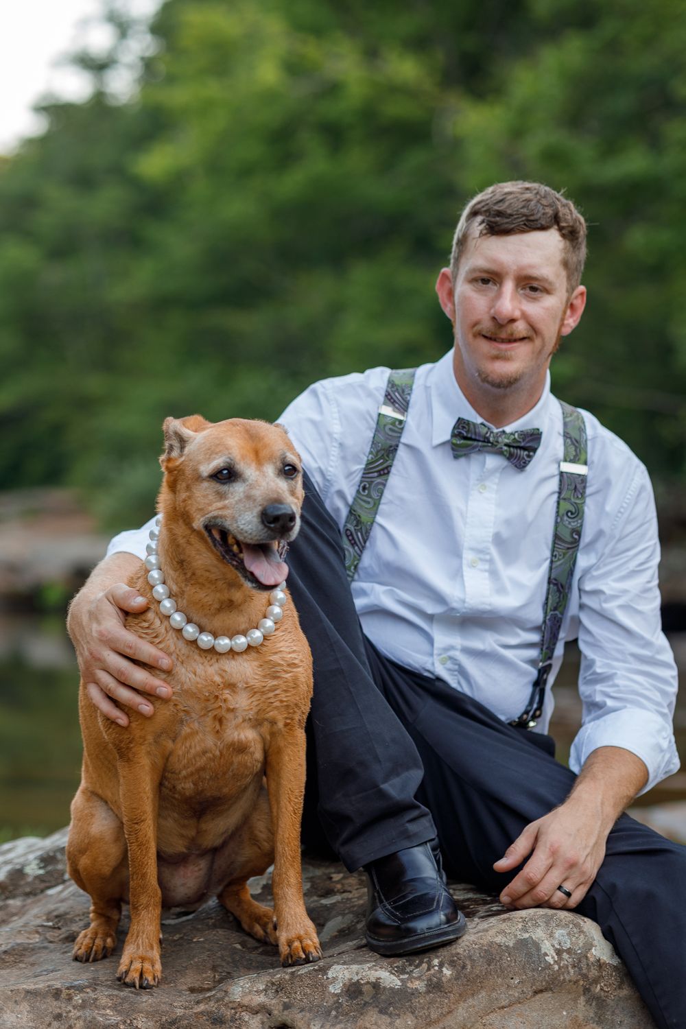 Person in formal attire poses with brown dog wearing pearl necklace on rocky outdoor setting.